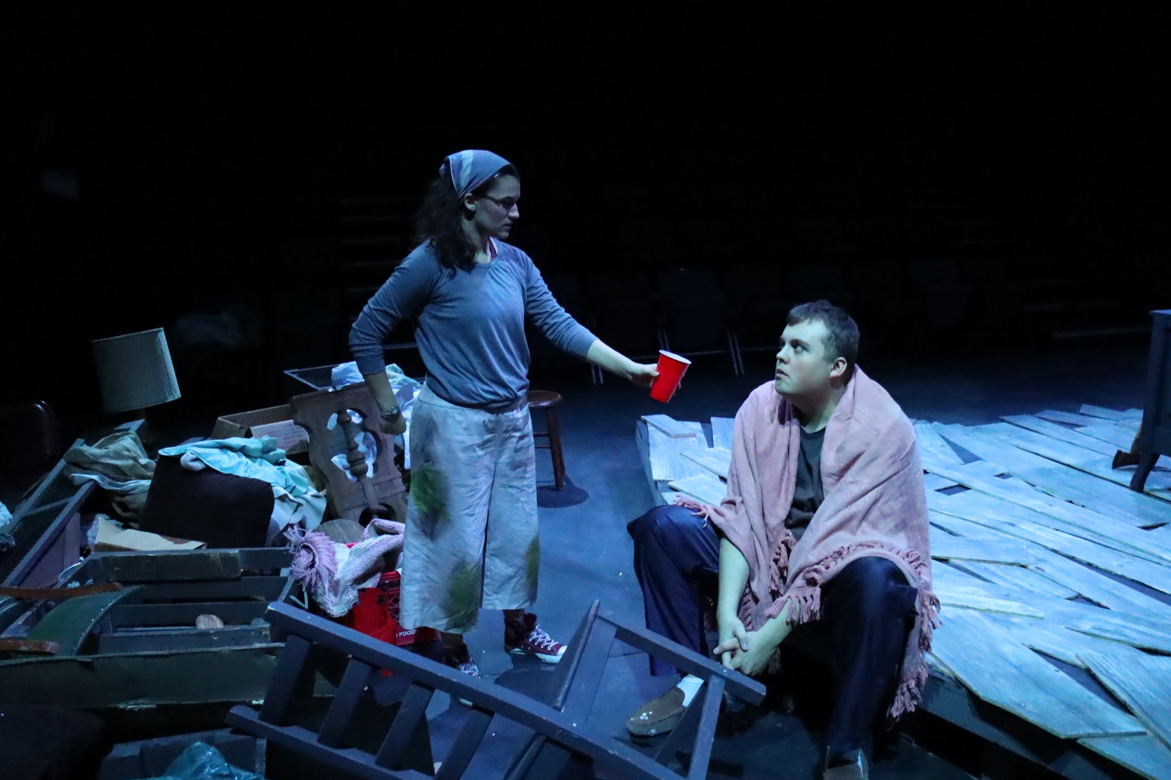 Two people on stage, one standing with a red cup, the other sitting, surrounded by scattered items and wooden planks.