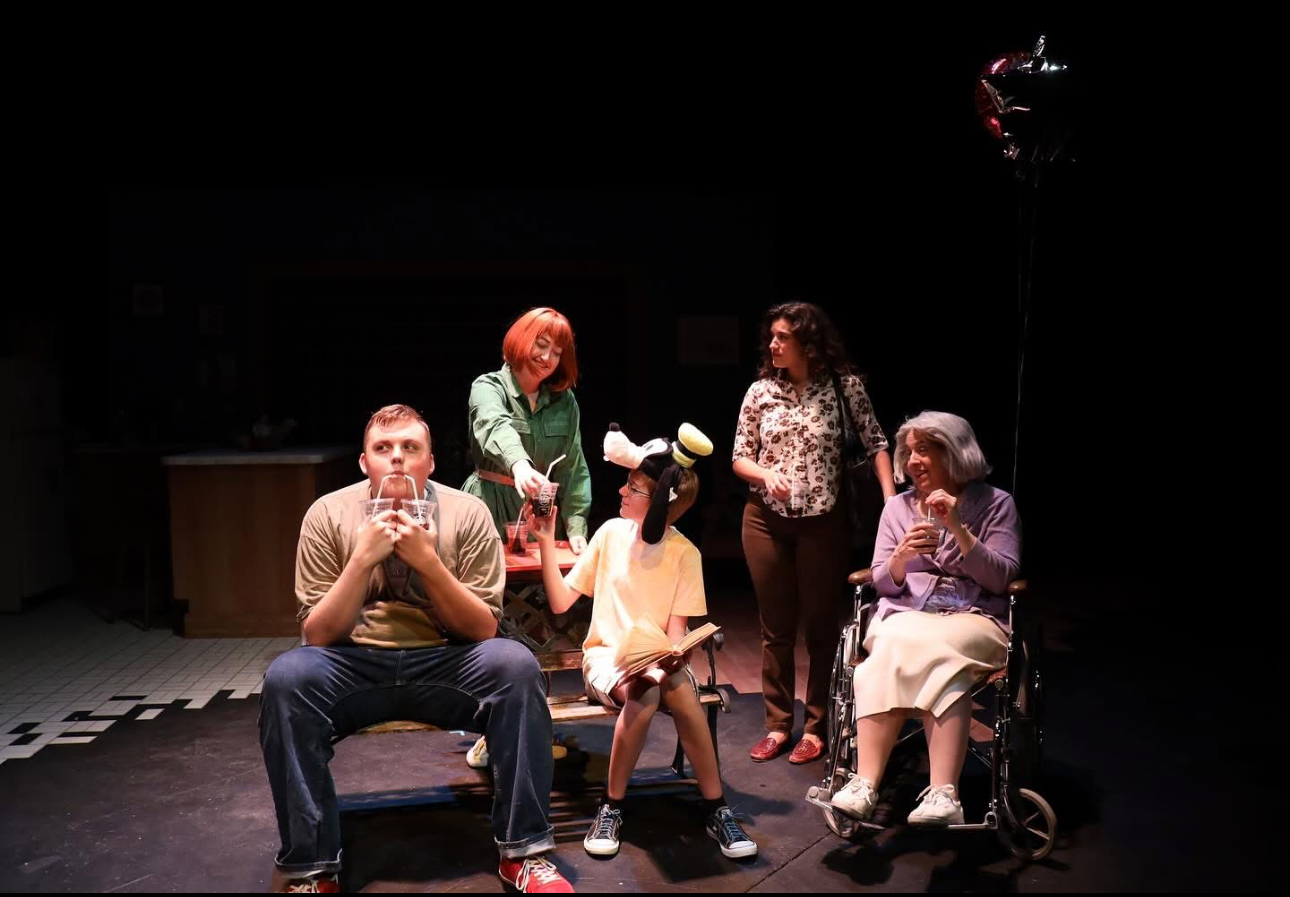 Five people, including children, are on a dark stage with minimal lighting, some sitting and some standing, engaged in a performance.