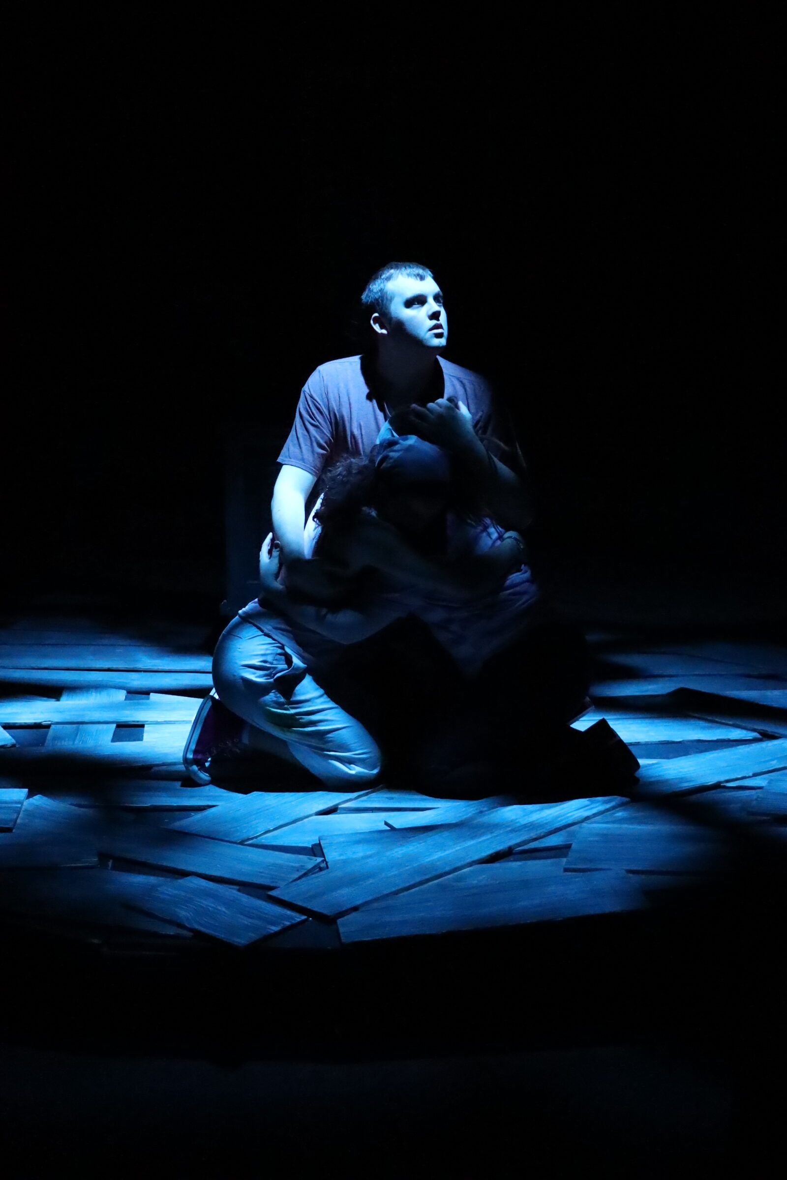 Person sitting on a wooden floor in a dark room, illuminated by blue light, with a contemplative expression.