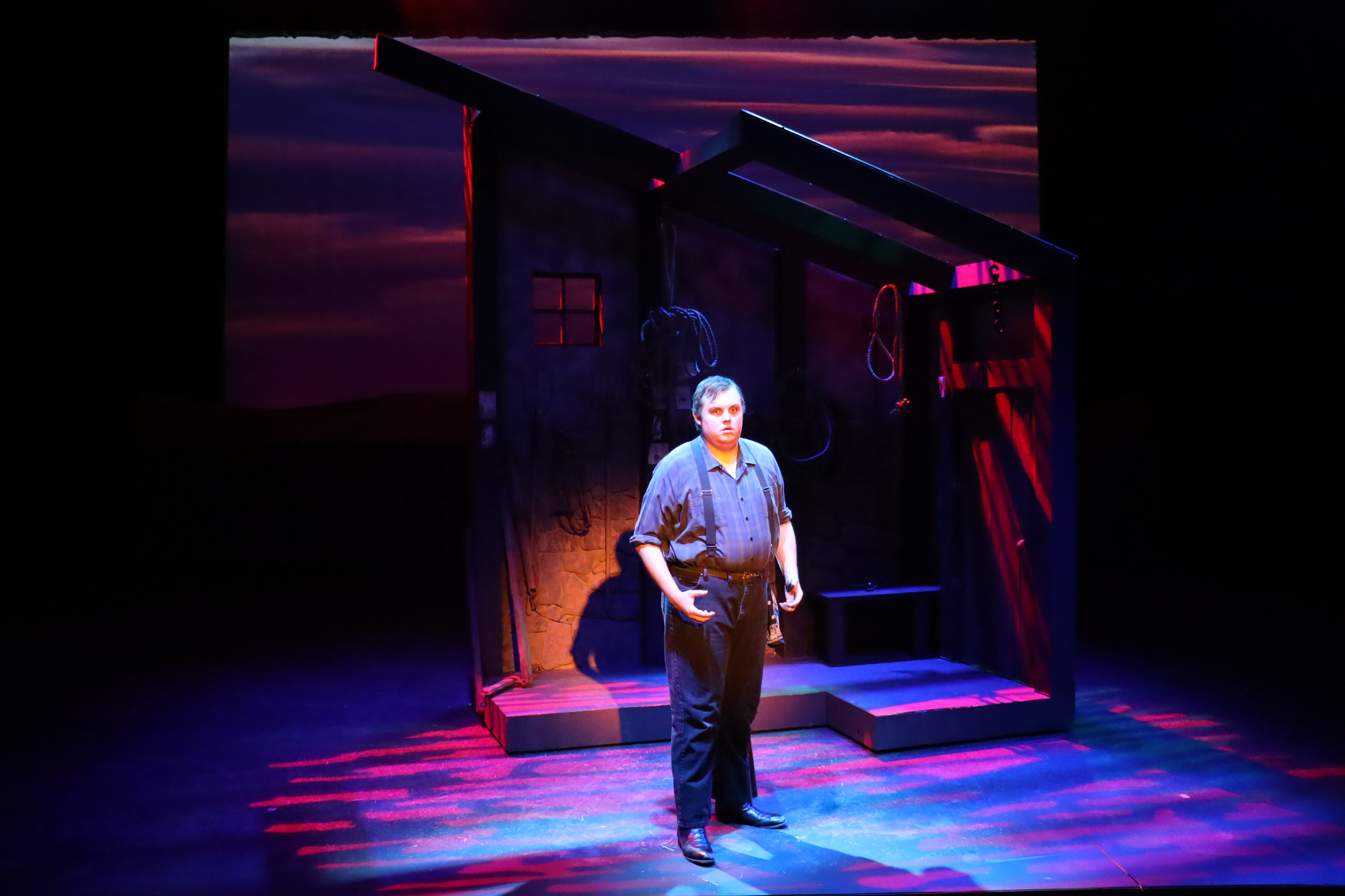 Person standing on stage in front of a small house set, with colorful lighting and a dark background.