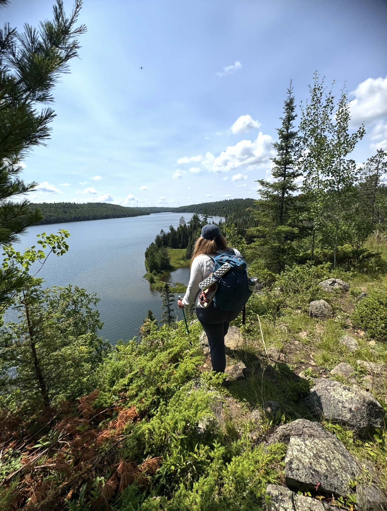 Person hiking on a trail beside a lake with trees and hills in the background under a blue sky with clouds.
