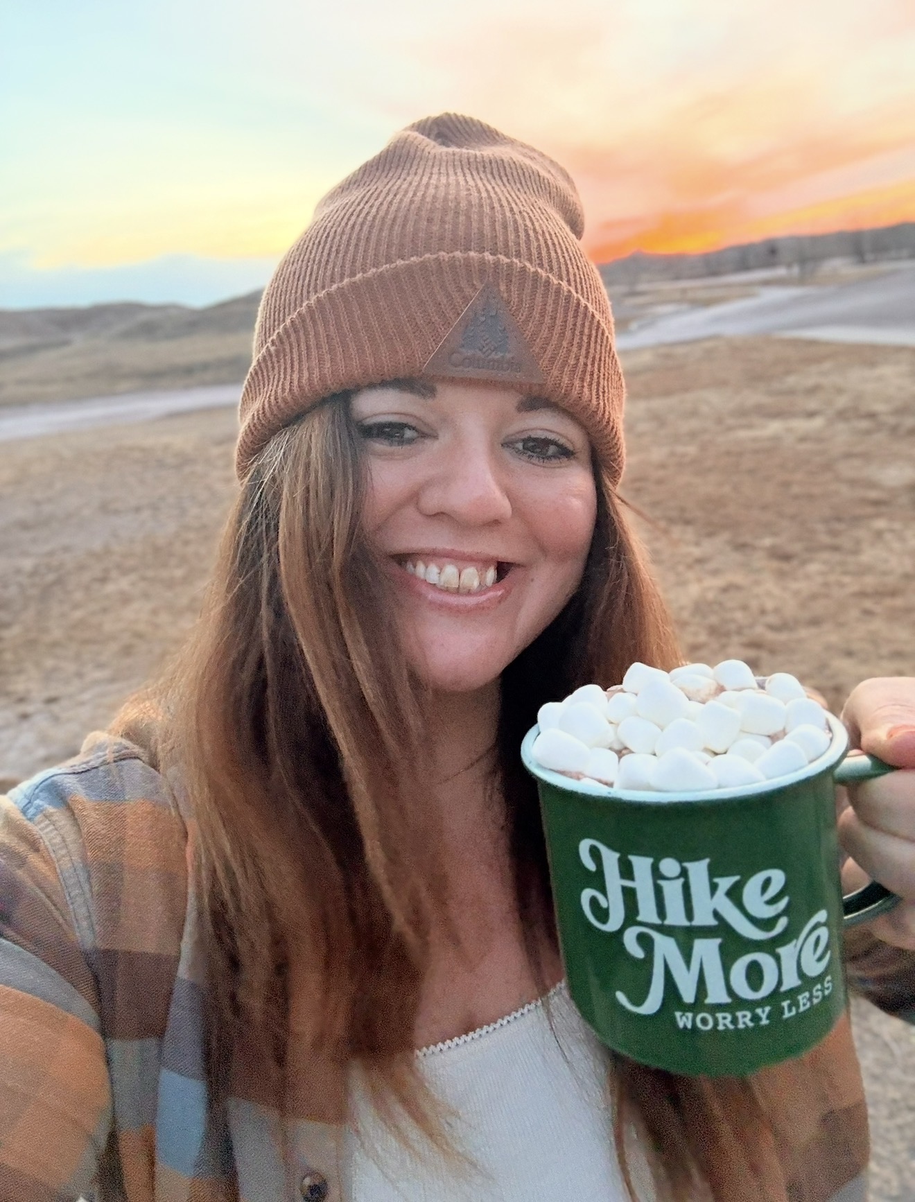 Woman smiling outdoors holding a mug with marshmallows, wearing a brown beanie and plaid shirt, sunset in background.