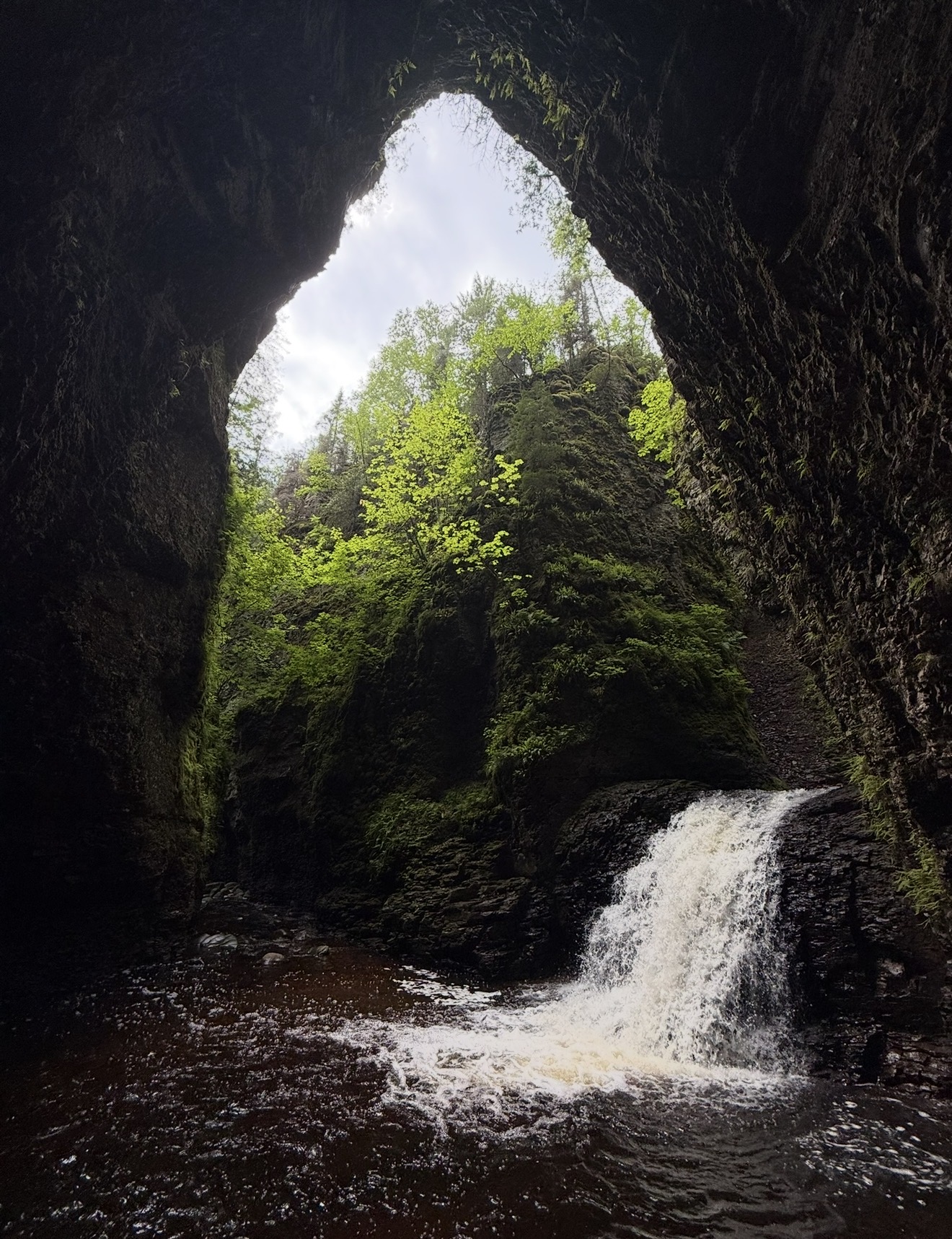 View through a cave opening showing a waterfall, lush green trees, and a cloudy sky.