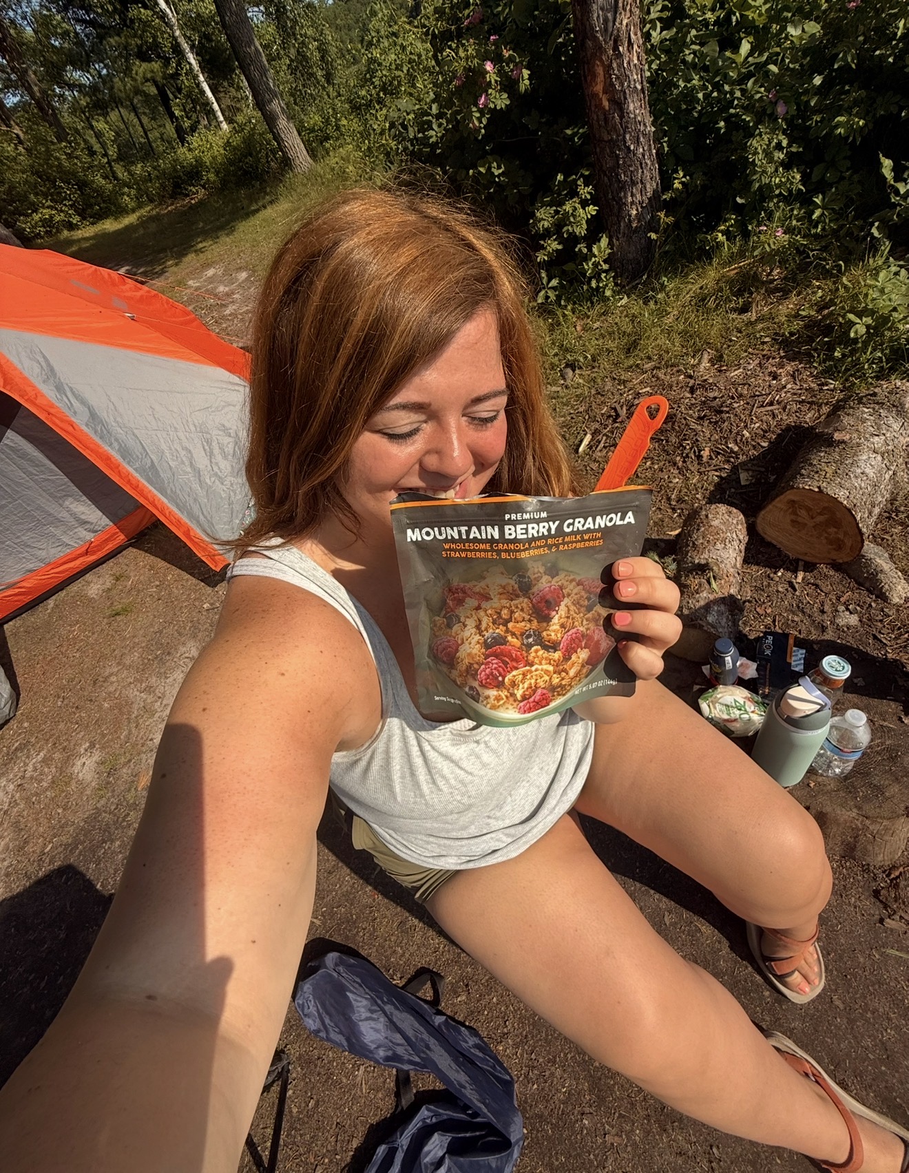 Person sitting outdoors near a campfire holding a Mountain Berry Granola packet, with an orange and gray tent nearby.