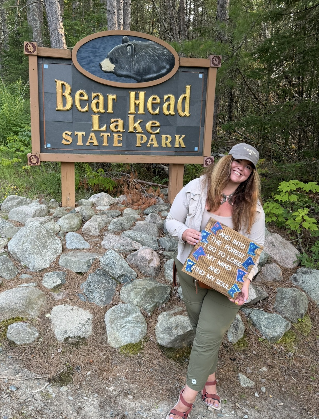 Woman holding a sign in front of a Bear Head Lake State Park sign, surrounded by rocks and trees.