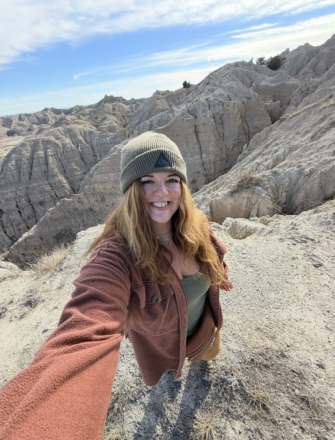 Woman smiling in a desert landscape with rocky formations, wearing a beanie and brown jacket.