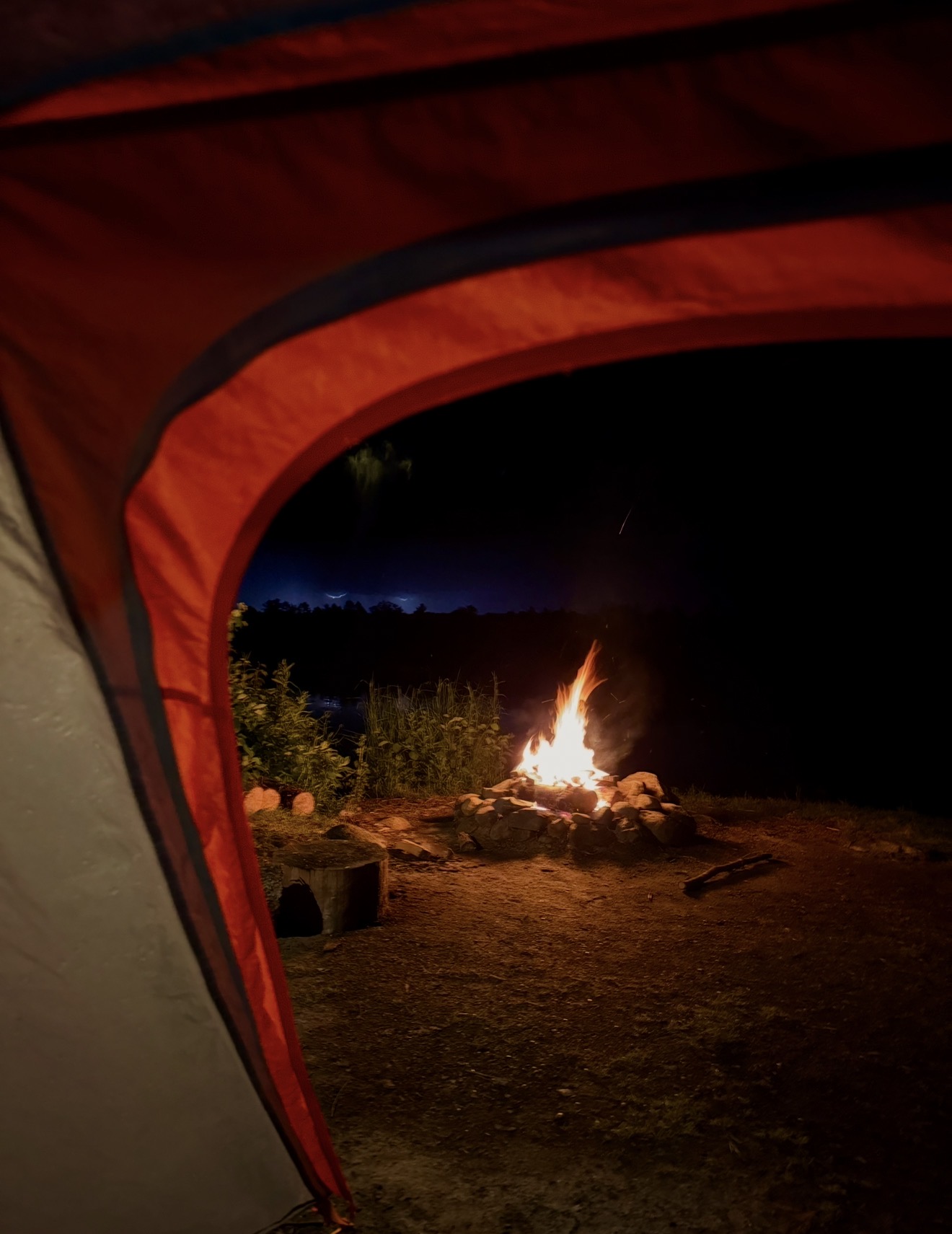 Campfire burning outdoors at night seen through a tent opening, with trees in the background.