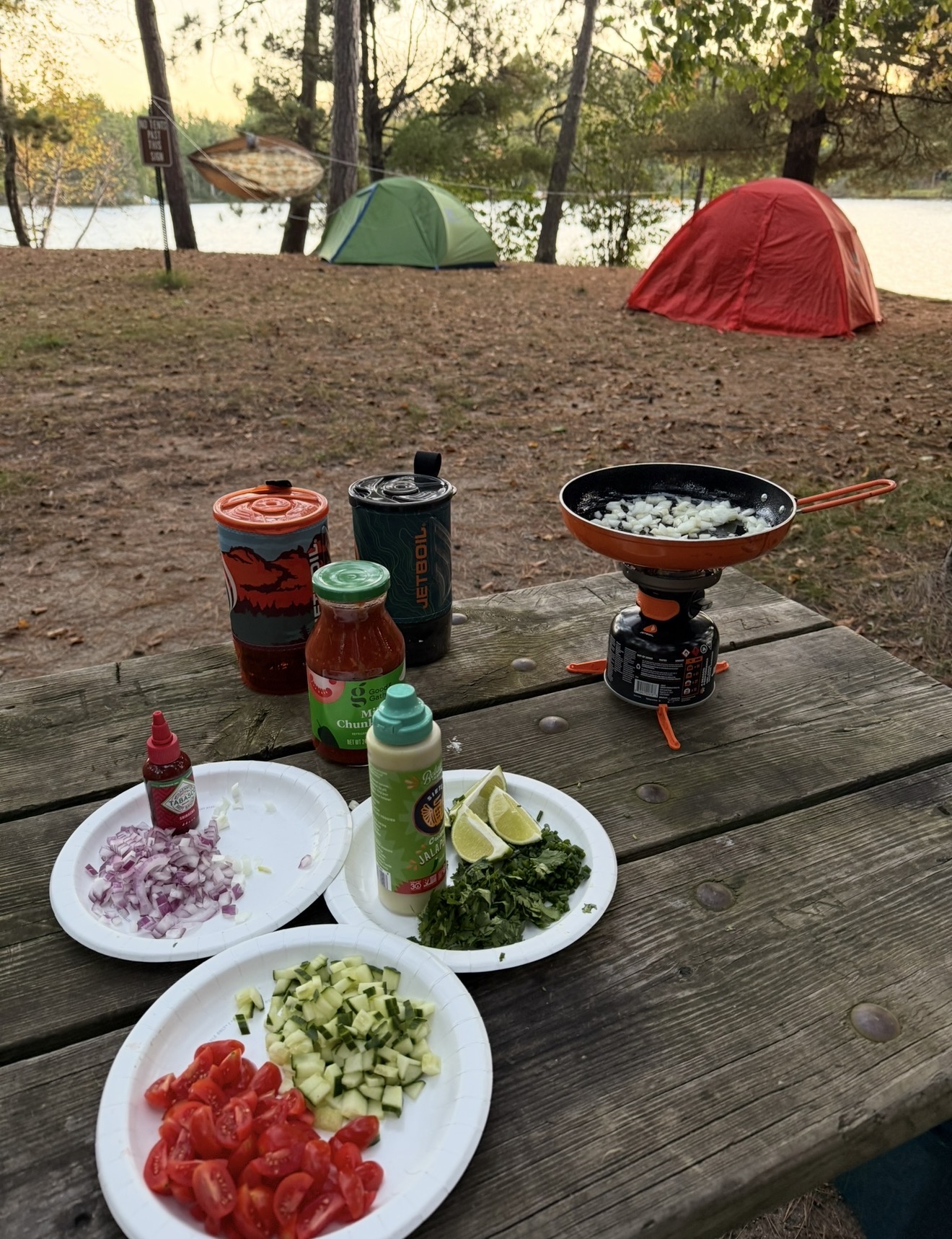 Camping table with food and a portable stove, two tents in the background near a river, trees, and outdoor setting.