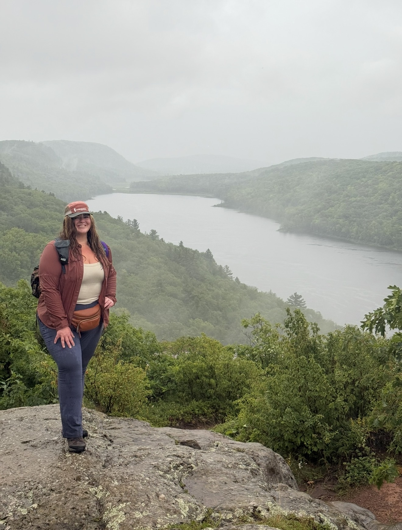 Woman standing on a rocky ledge overlooking a river and green hills under cloudy sky.