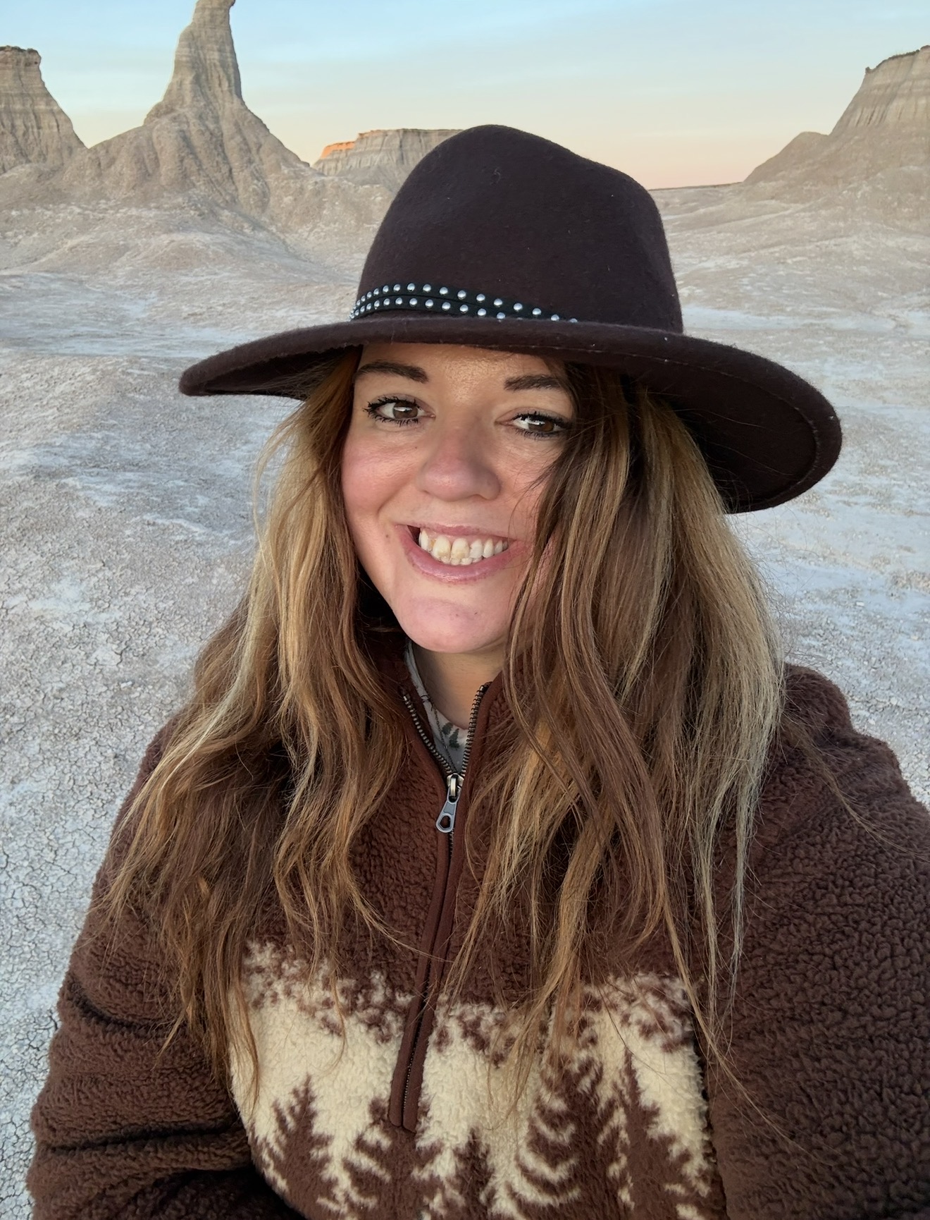 Young woman with long hair smiling, wearing a black hat and brown jacket, in a desert landscape with rock formations.
