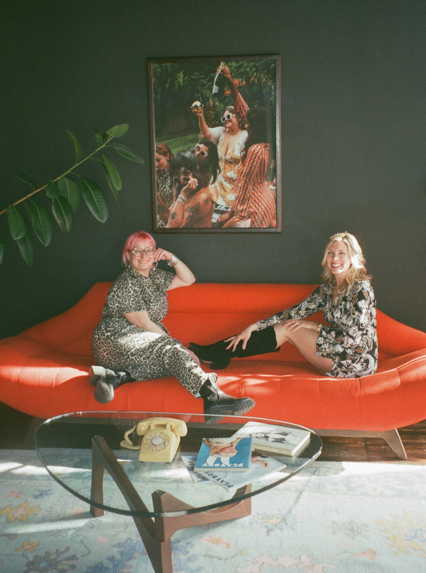Two women sit on an orange sofa in a room with dark green walls, a framed picture, and a glass coffee table.