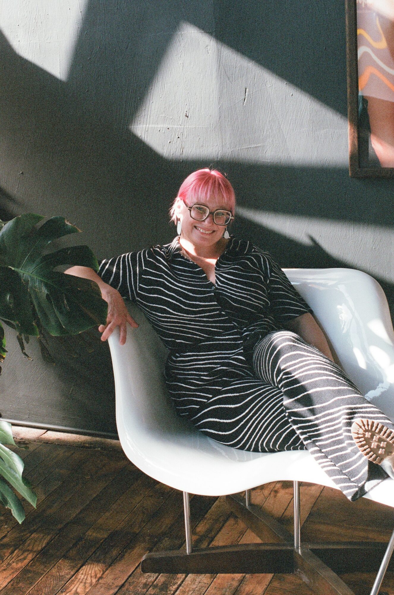 Person with pink hair and glasses sitting on a white chair, wearing a black and white striped dress, in a modern indoor space.