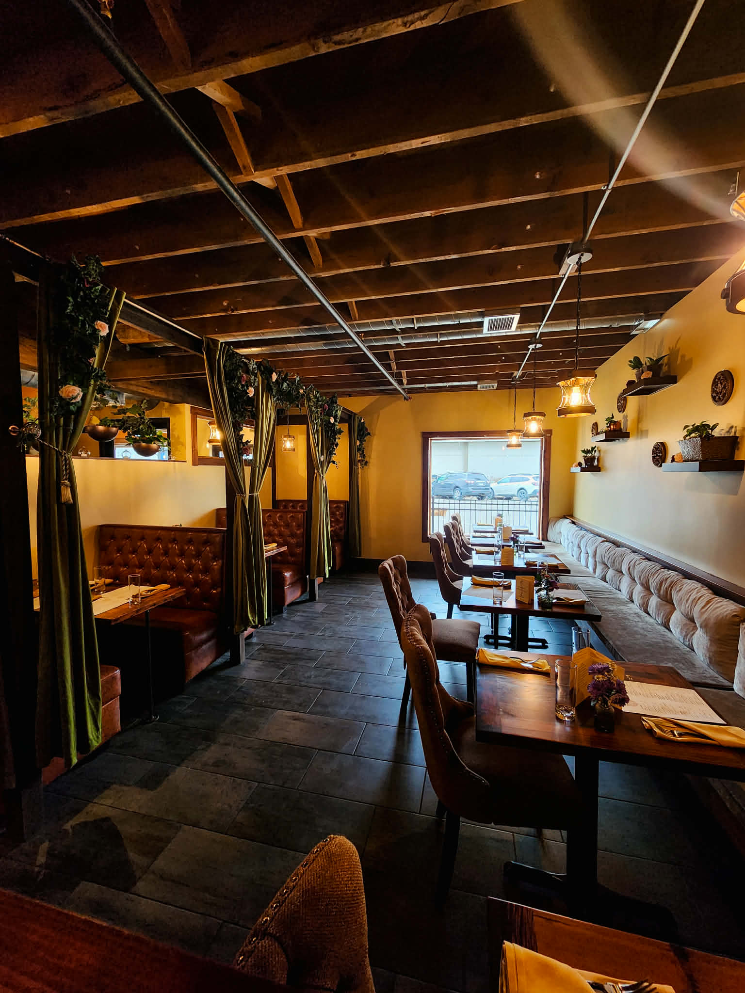 Restaurant interior with wooden ceiling, booth seating, and a window view, decorated with plants and shelves, dim lighting.