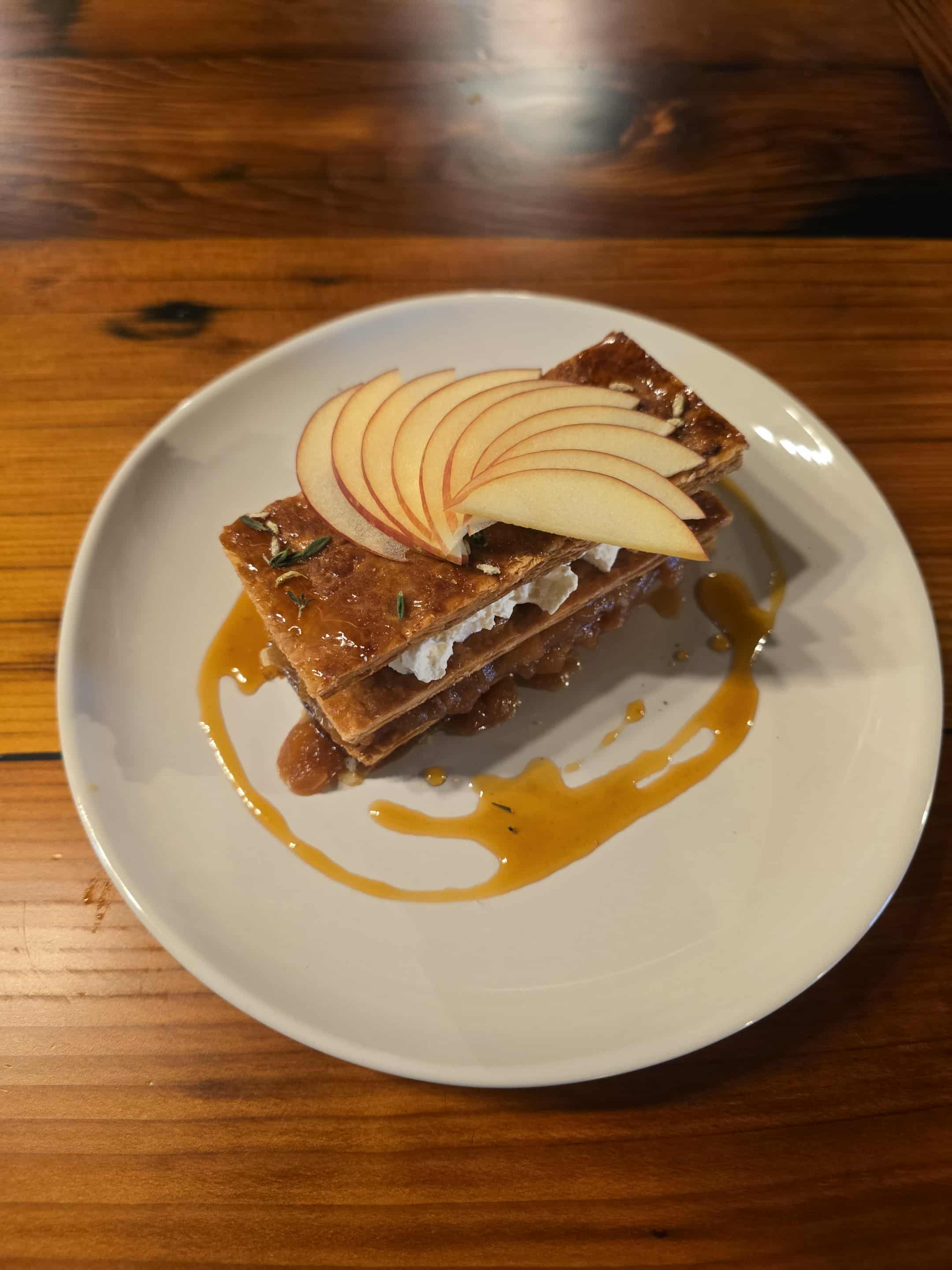 Stacked waffles topped with sliced apples and caramel sauce on a white plate, on a wooden table.