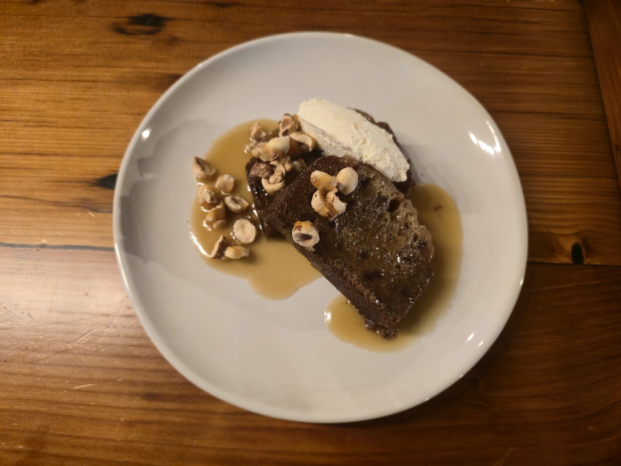 Chocolate cake with nuts, whipped cream, and caramel sauce on a white plate, on a wooden table.