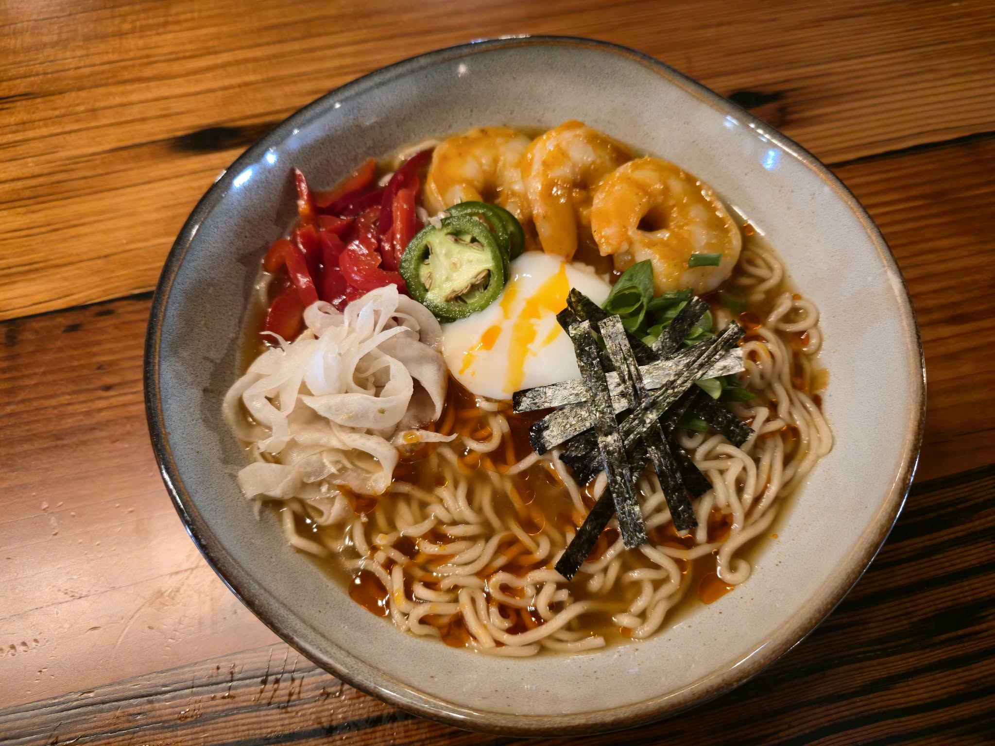 Bowl of ramen with noodles, sliced jalapeños, red peppers, seaweed, and a soft-boiled egg on a wooden table.