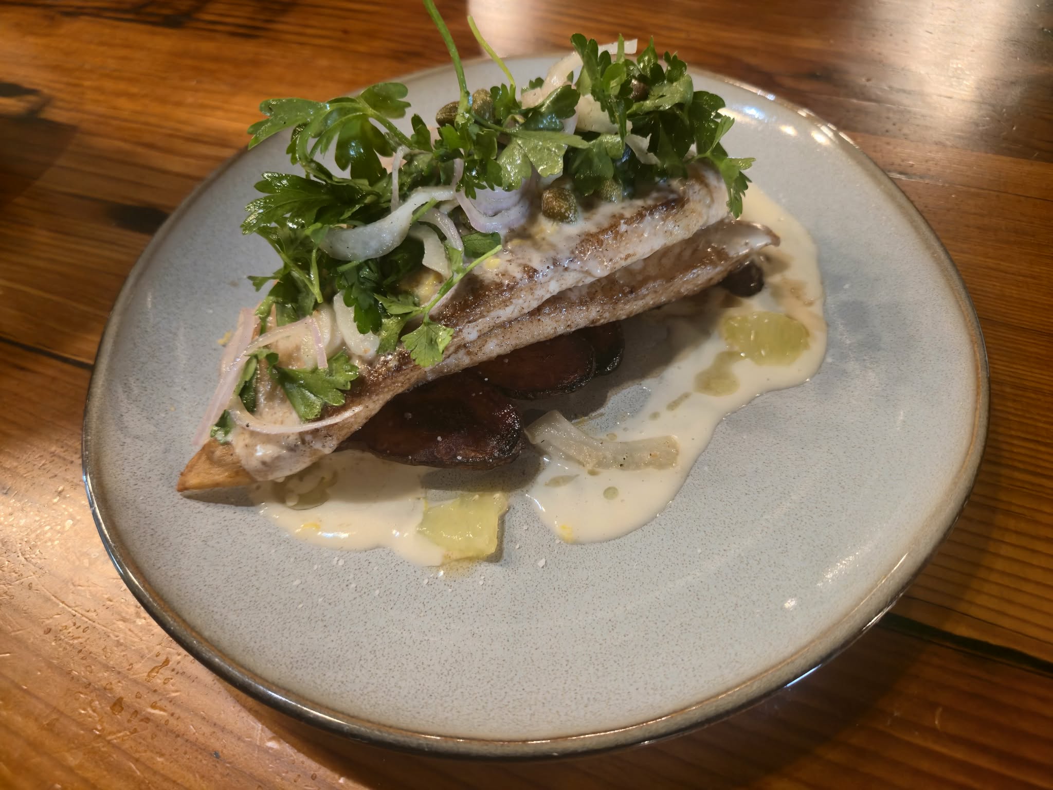 A plated dish with fish, greens, and sauce on a light-colored plate, on a wooden table.