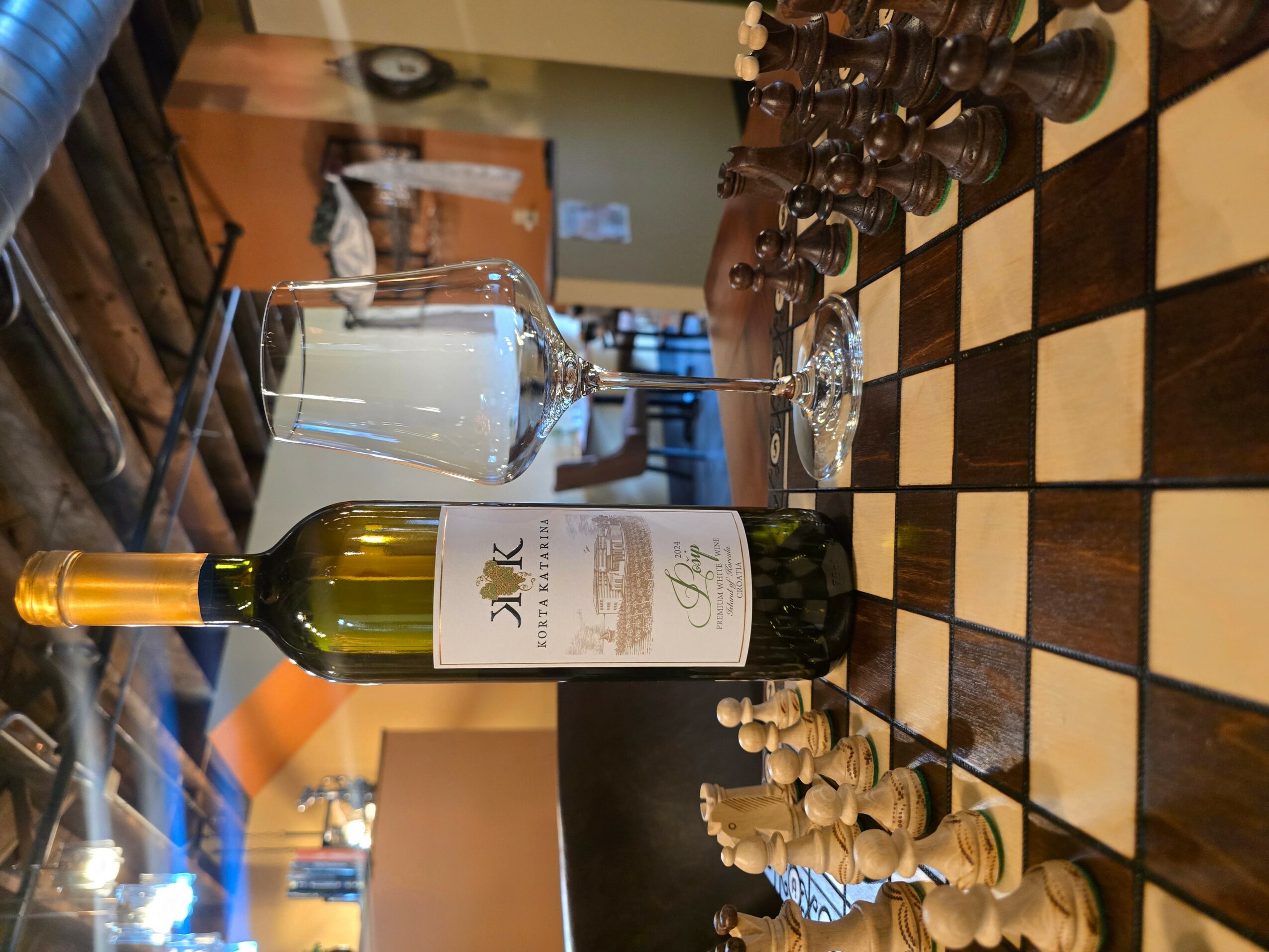 A bottle of white wine, a glass of white wine, and a chessboard with chess pieces on a table.