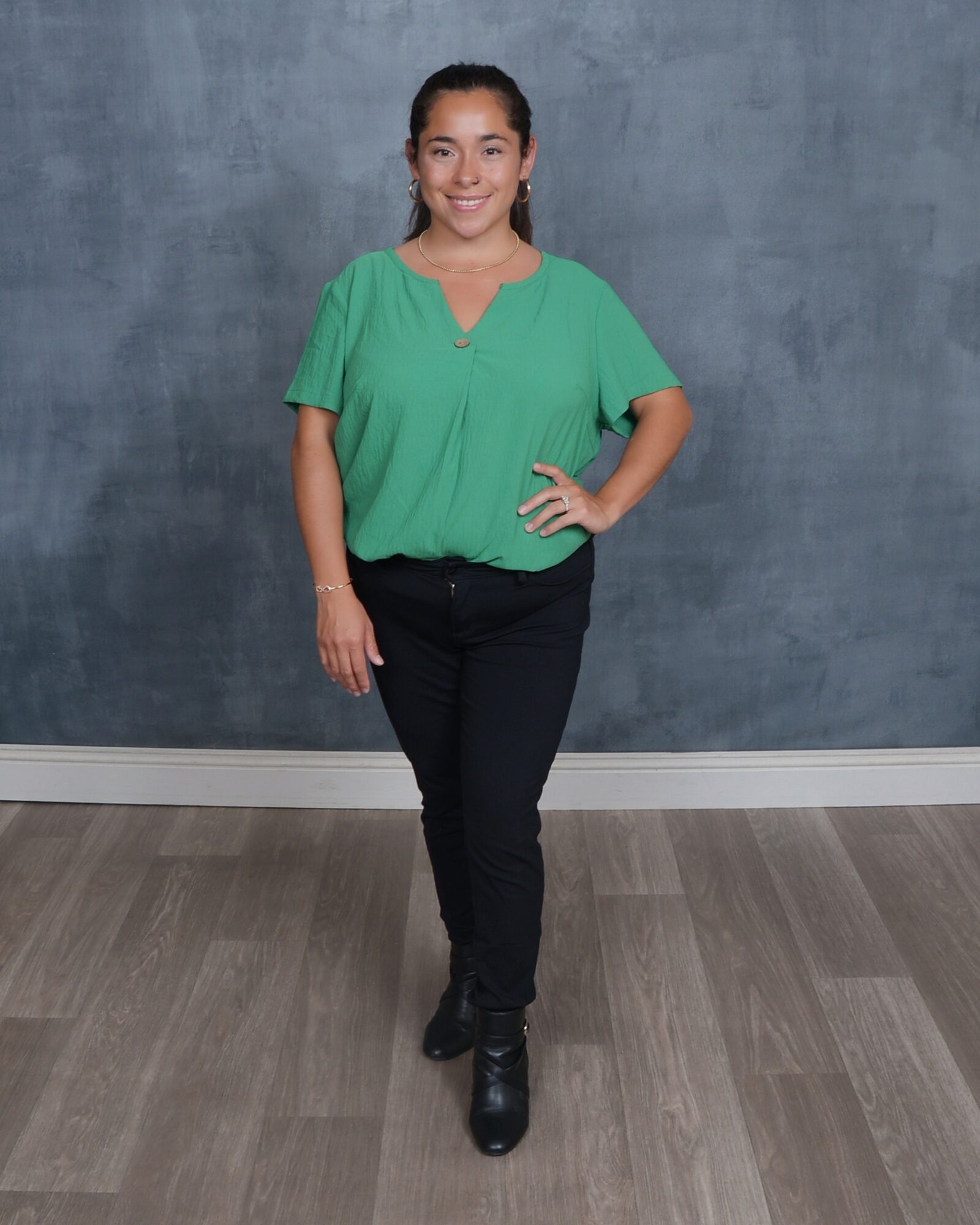Young woman standing with hand on hip, smiling, wearing green blouse and black pants, against a gray wall background.