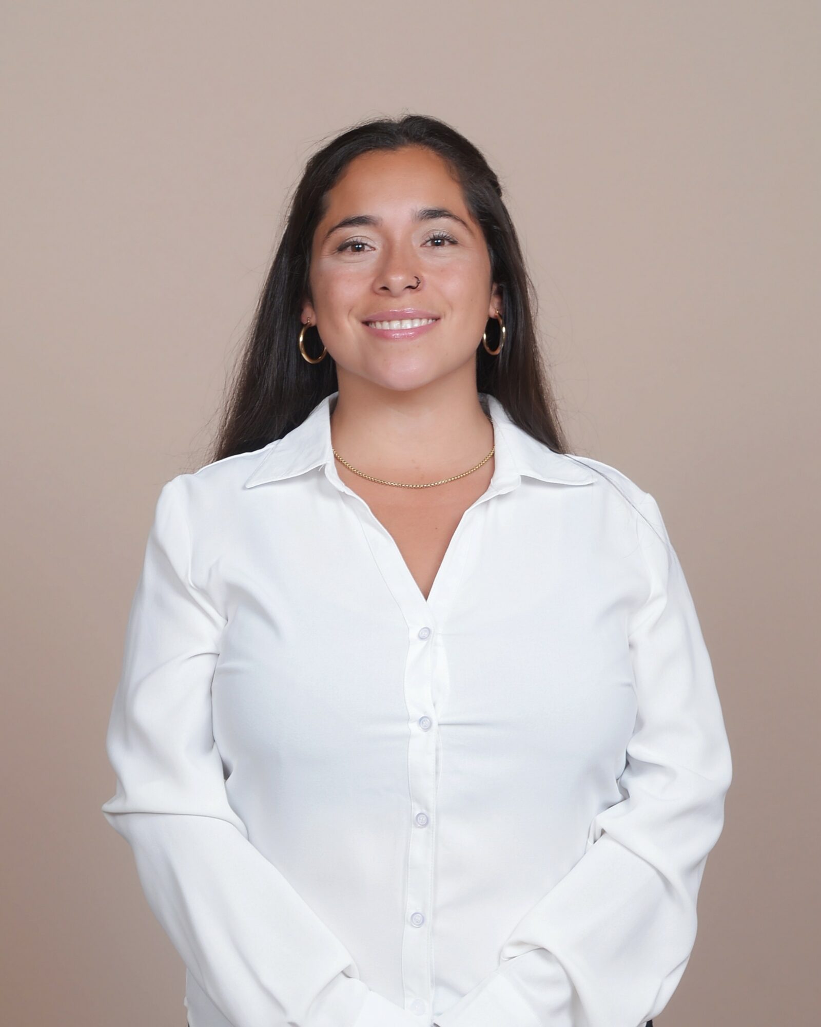 Young woman with long dark hair, wearing a white button-up shirt and hoop earrings, smiling against a beige background.