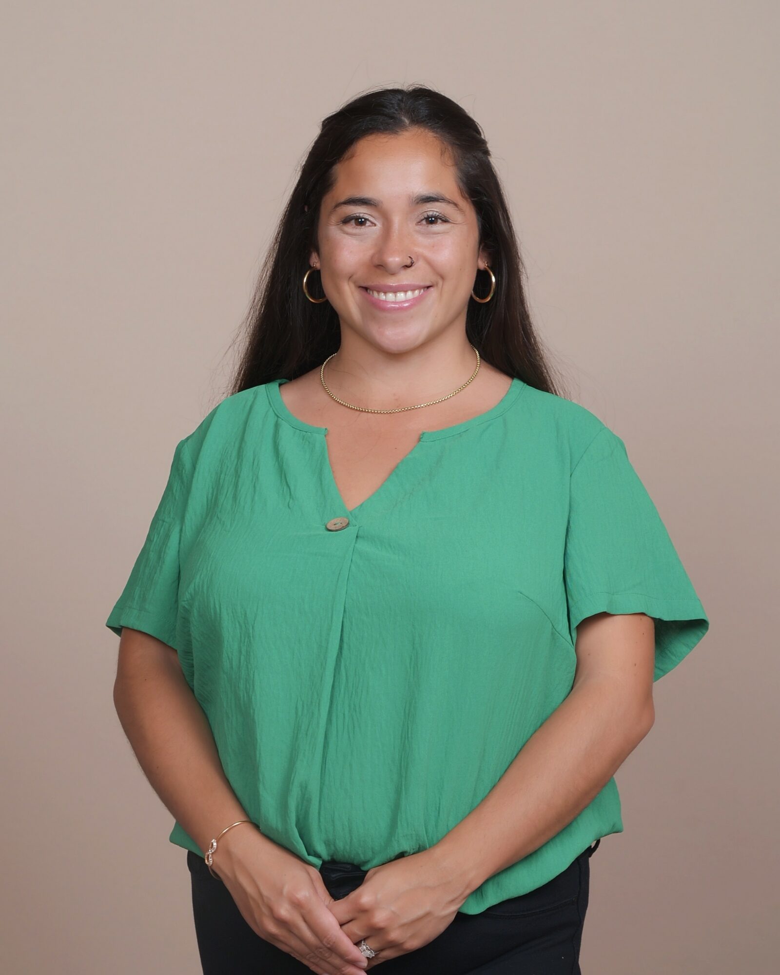 Smiling woman with long dark hair, wearing a green blouse and hoop earrings, standing against a beige background.