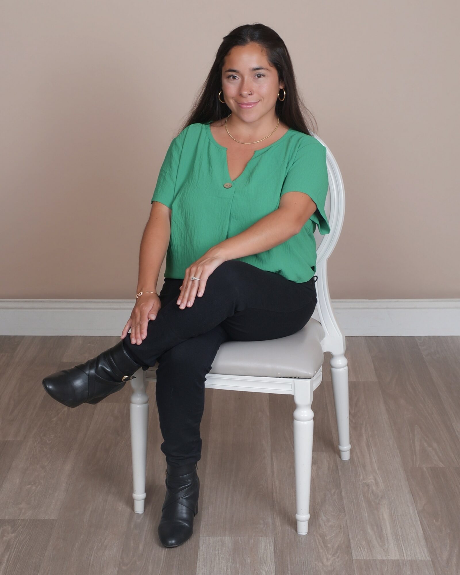 Young woman with long dark hair sitting on a white chair, wearing a green top and black pants, smiling.