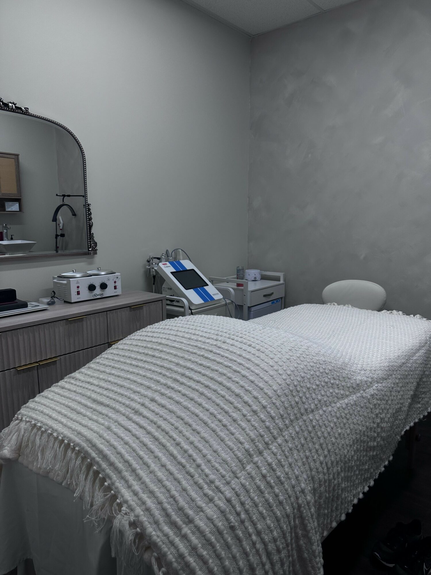 Hospital bed with a white blanket, bedside table with medical equipment, and a wall-mounted lamp.