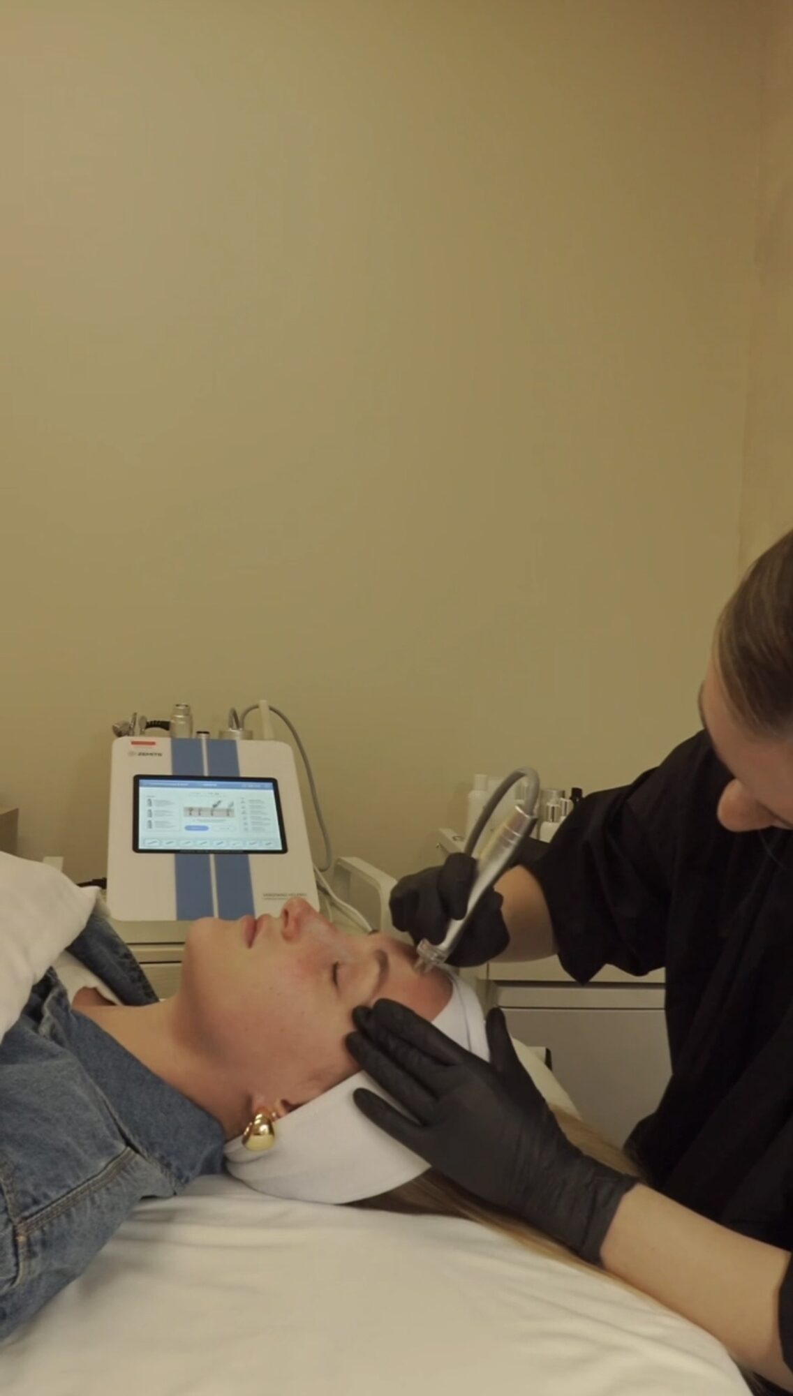Person receiving a facial treatment while lying on a bed, with a technician wearing black gloves performing the procedure.