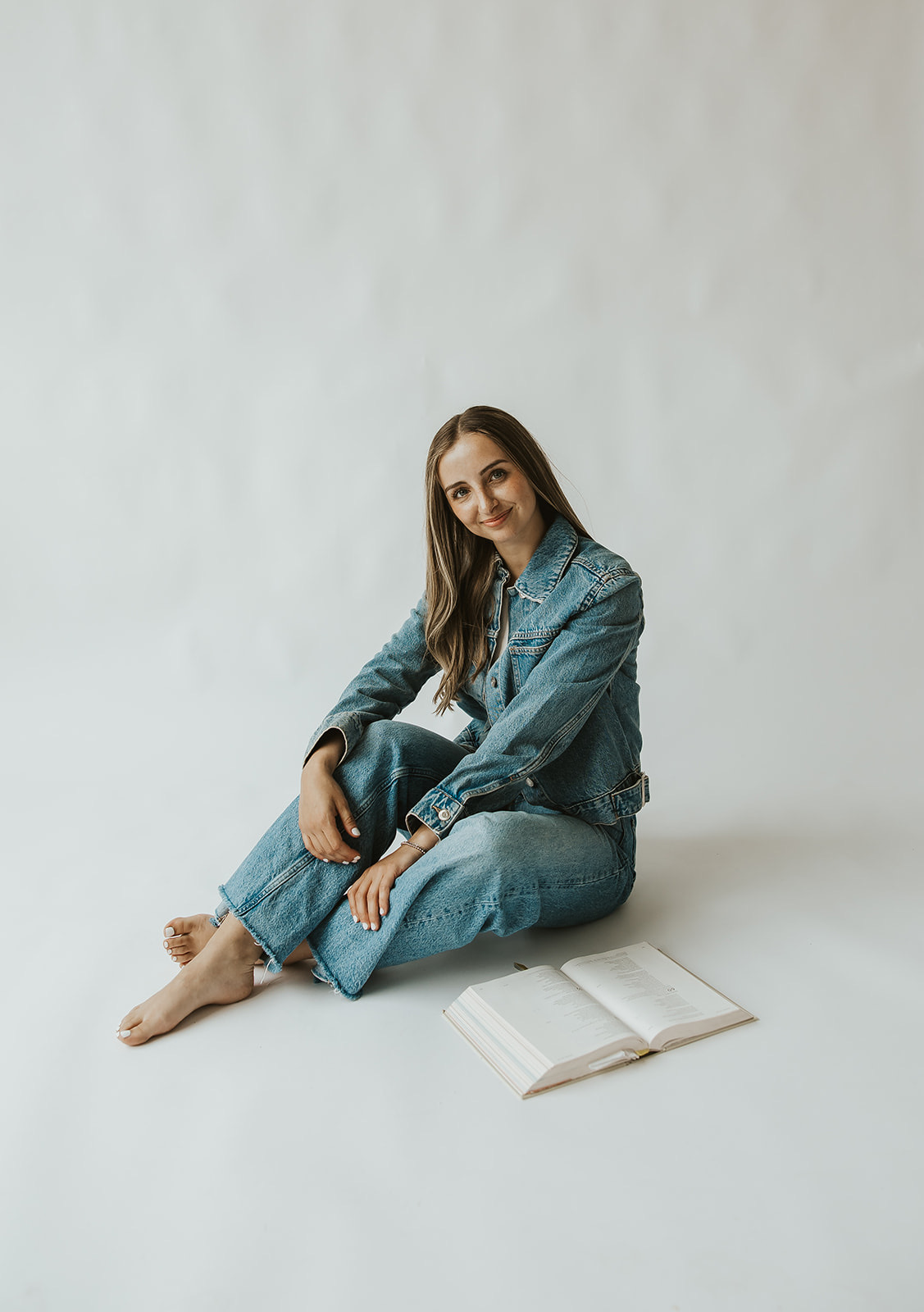 Young woman sitting on the floor with an open book nearby, wearing a denim jacket and jeans, against a plain background.