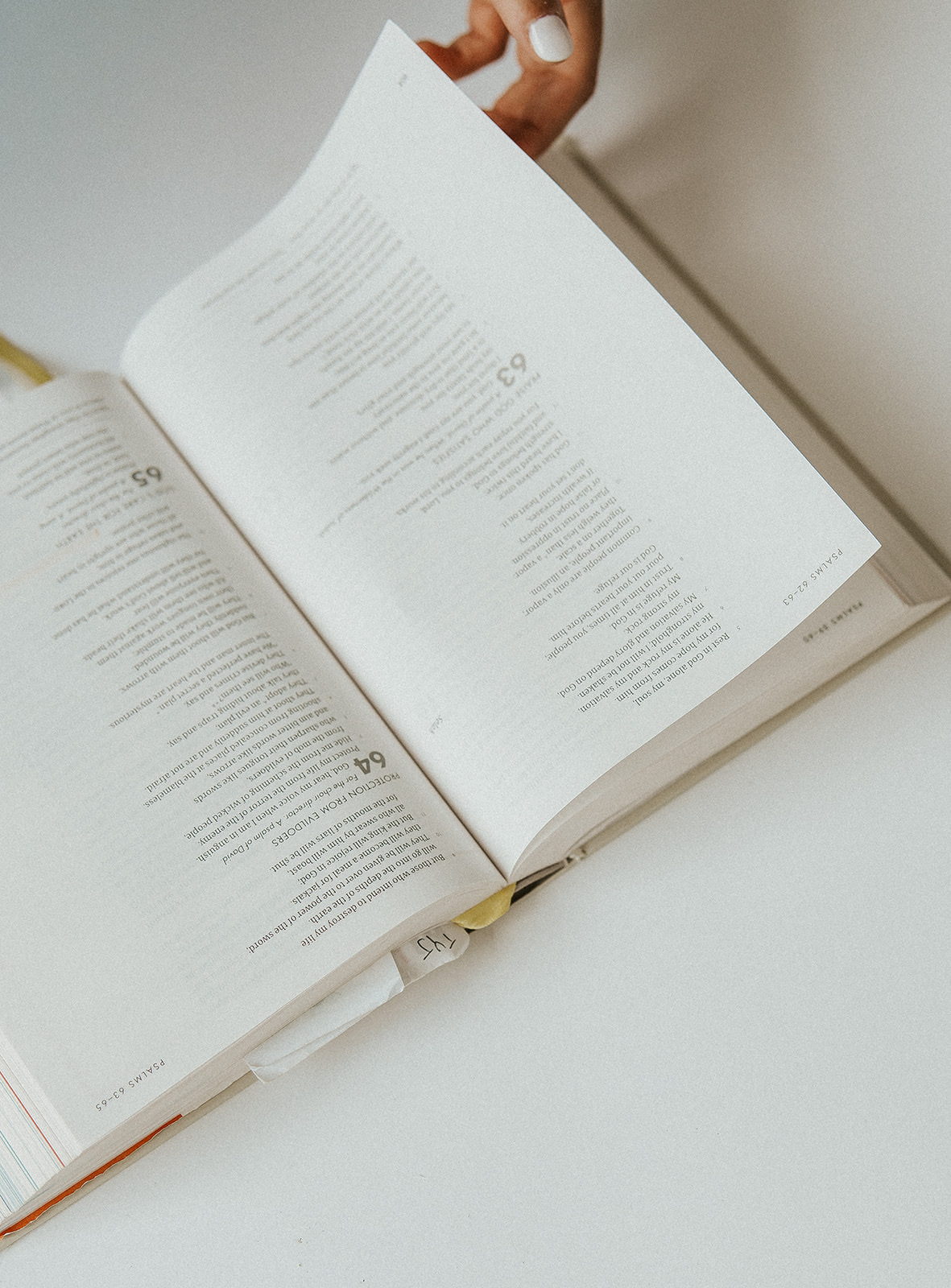 Person's hand holding open book with text, viewed from above, on a white surface.
