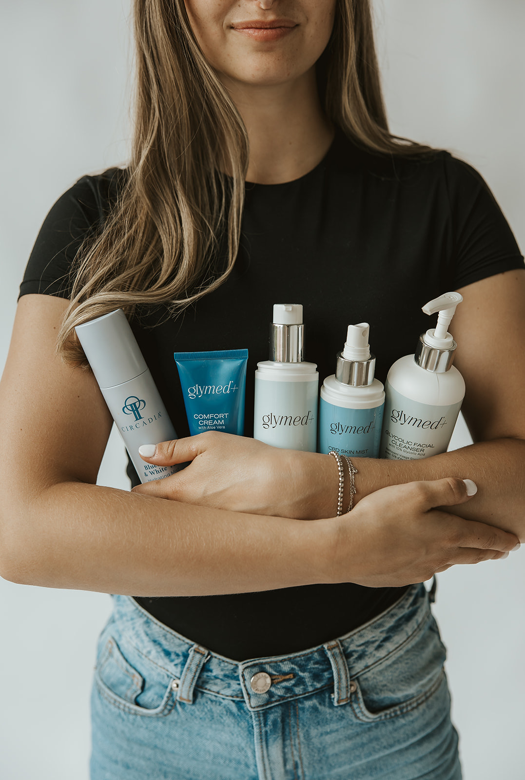 Woman holding skincare products, wearing a black shirt and jeans, with long hair and a bracelet, against a plain background.