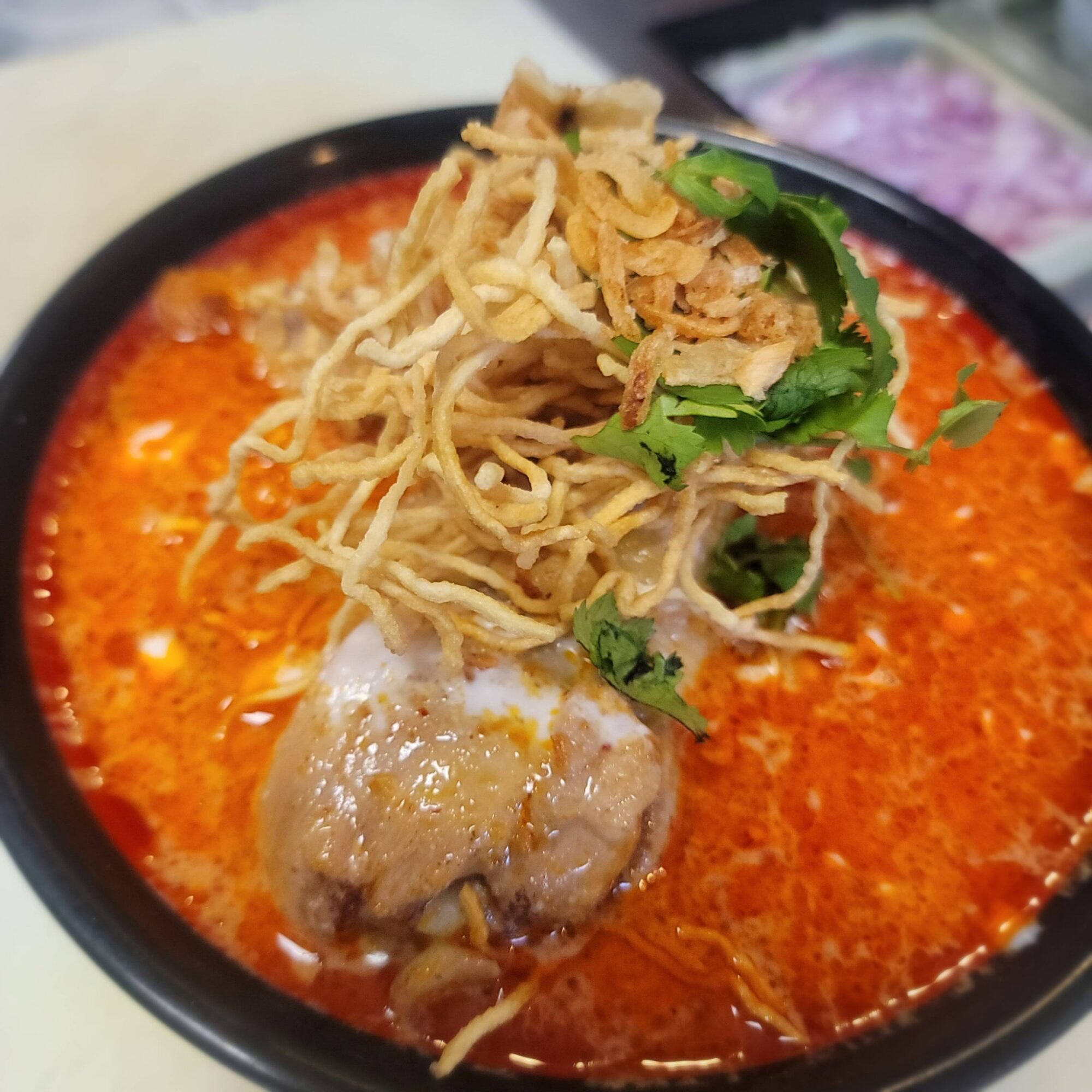 Bowl of spicy red soup with noodles, chicken, and herbs, topped with fried shallots and cilantro, close-up view.