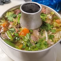 Bowl of noodle soup with vegetables, herbs, and meat, with a central cylindrical container, on a table.