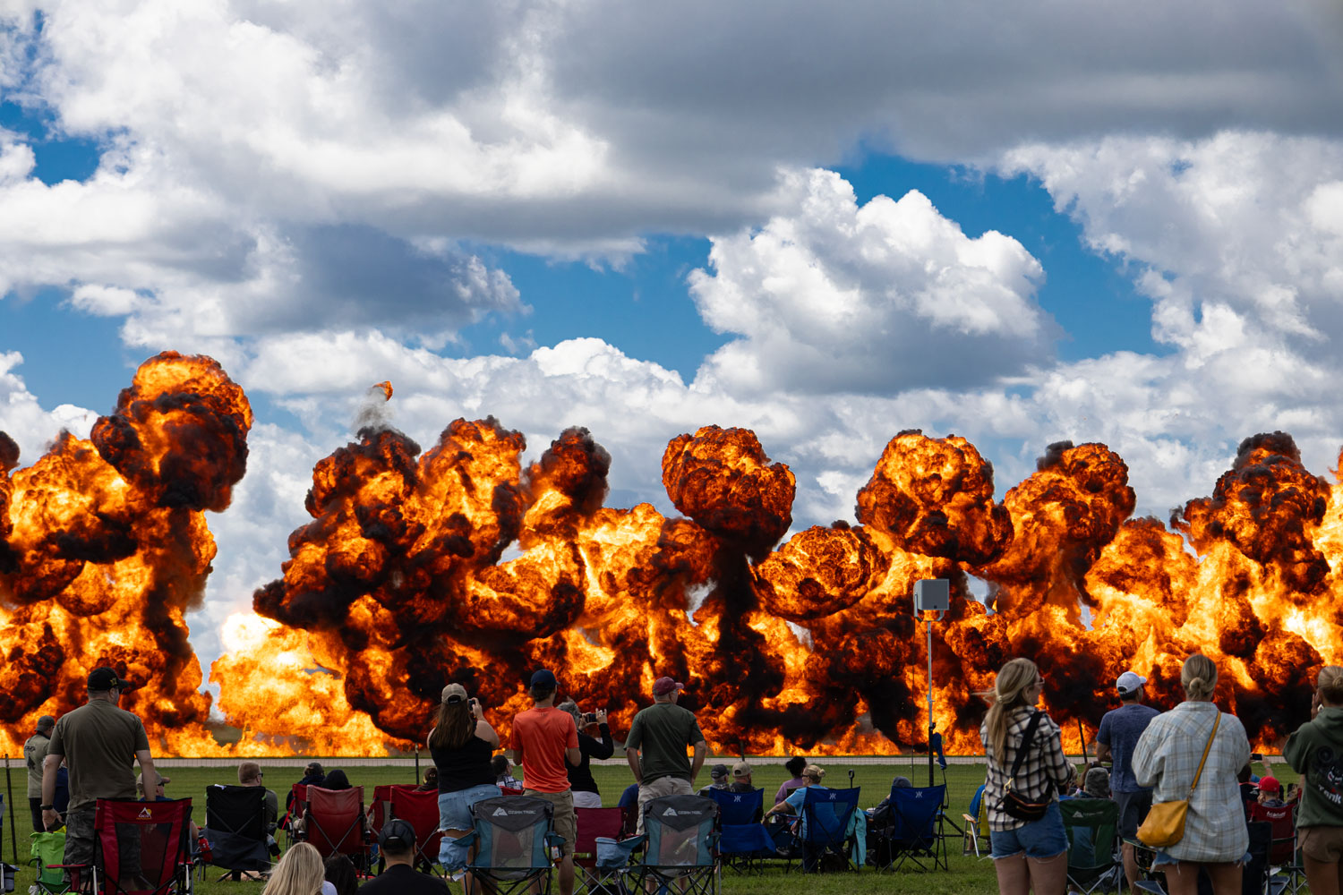 People watching an explosion with large fireballs and smoke in the sky, clouds overhead, and some seated on the ground.