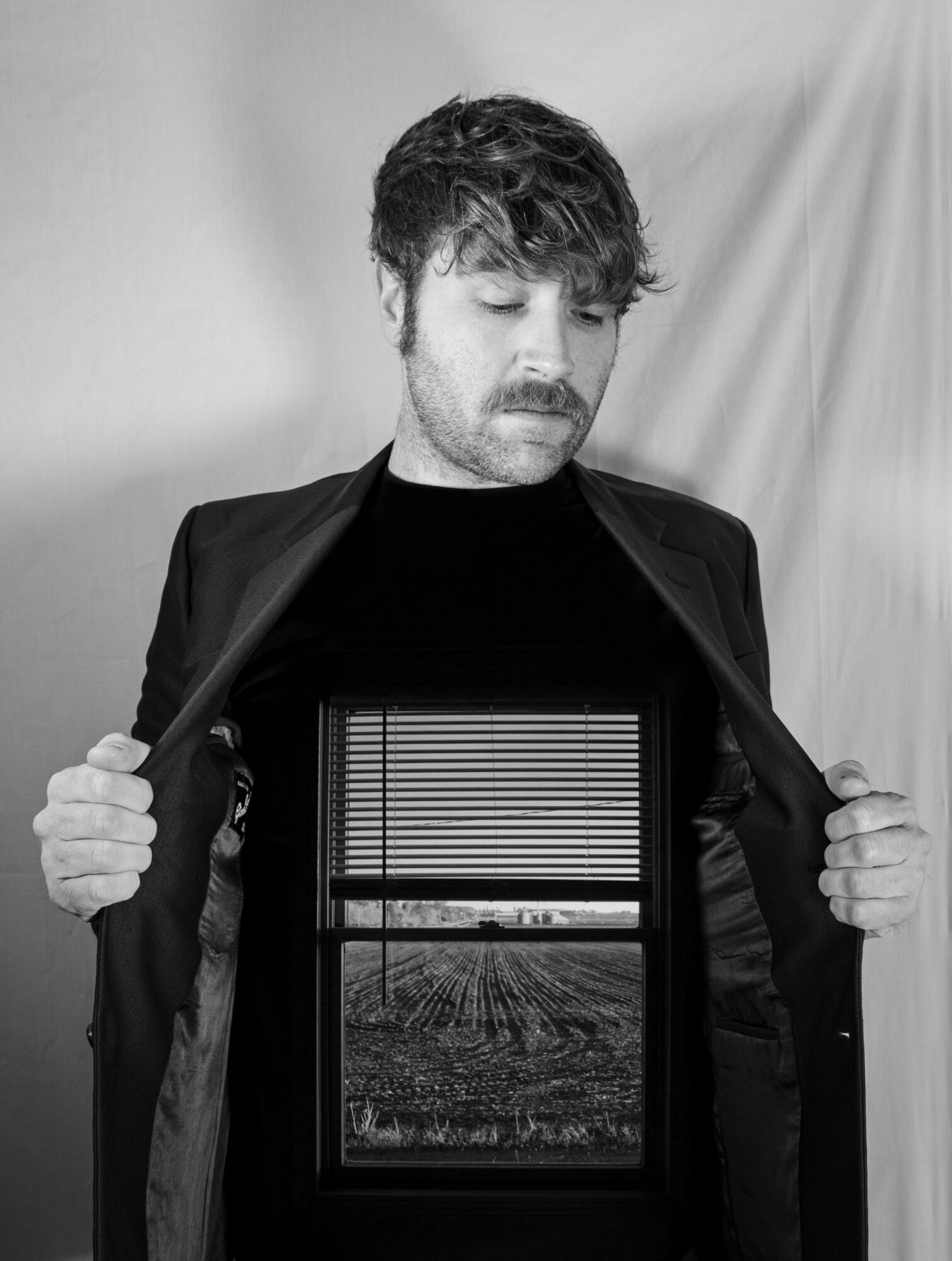 Man with tousled hair and beard opening a jacket to reveal a window with blinds and a landscape view inside.