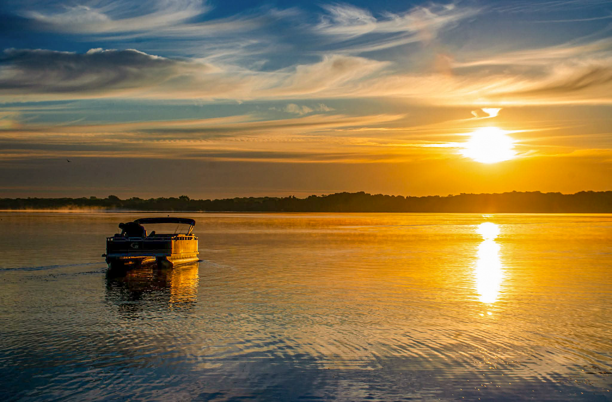 Sunset over a calm body of water with a boat, reflecting the sky's colors, and a distant shoreline.