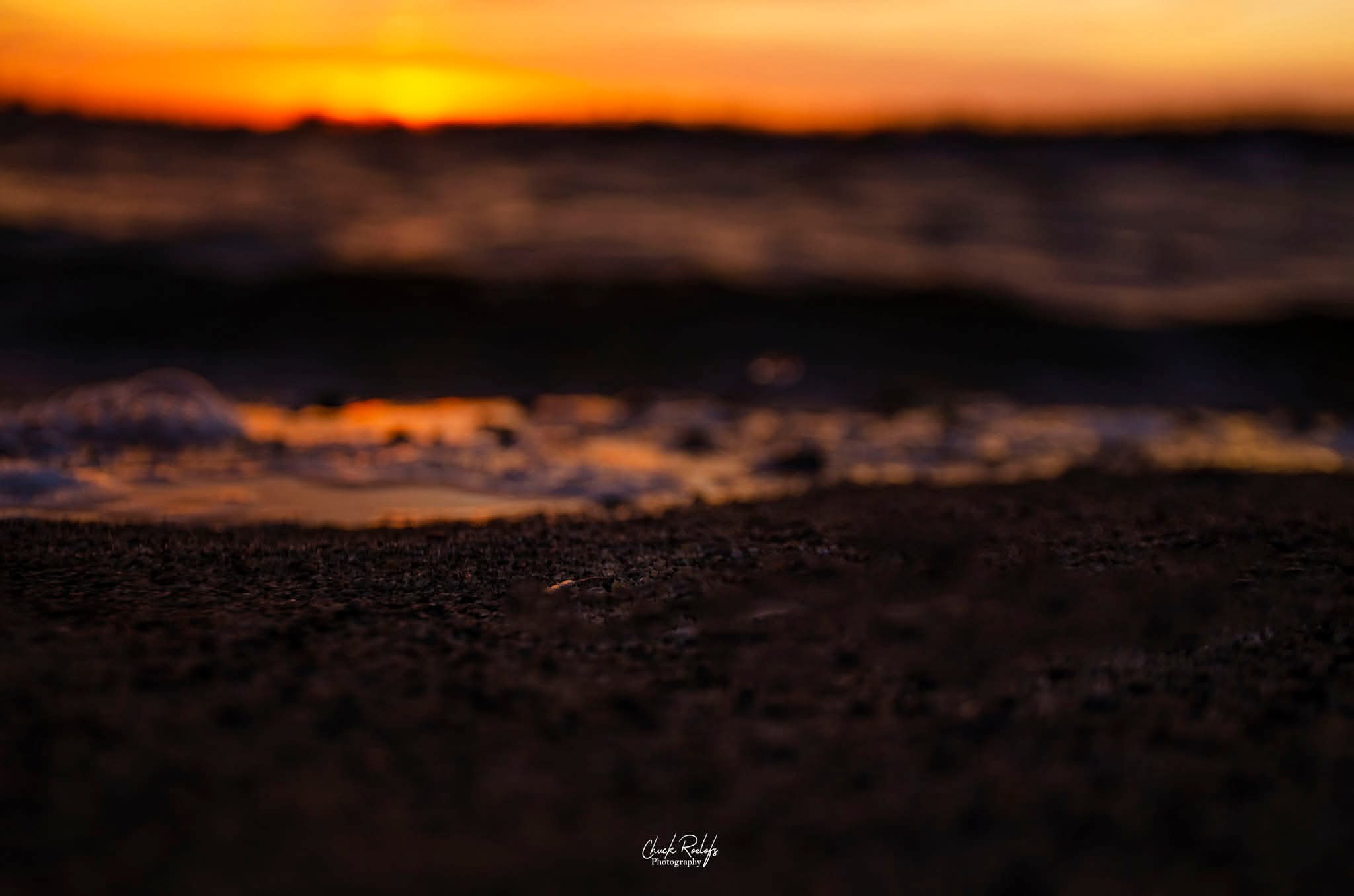 Sunset over the ocean with waves and dark sandy beach in foreground.