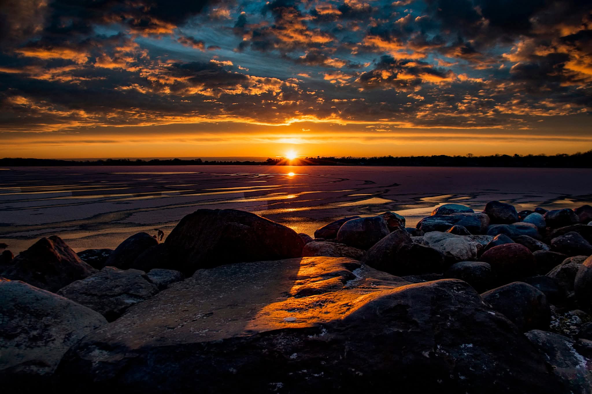 Sunset over a beach with rocks in the foreground and a sky filled with clouds, orange and dark hues.
