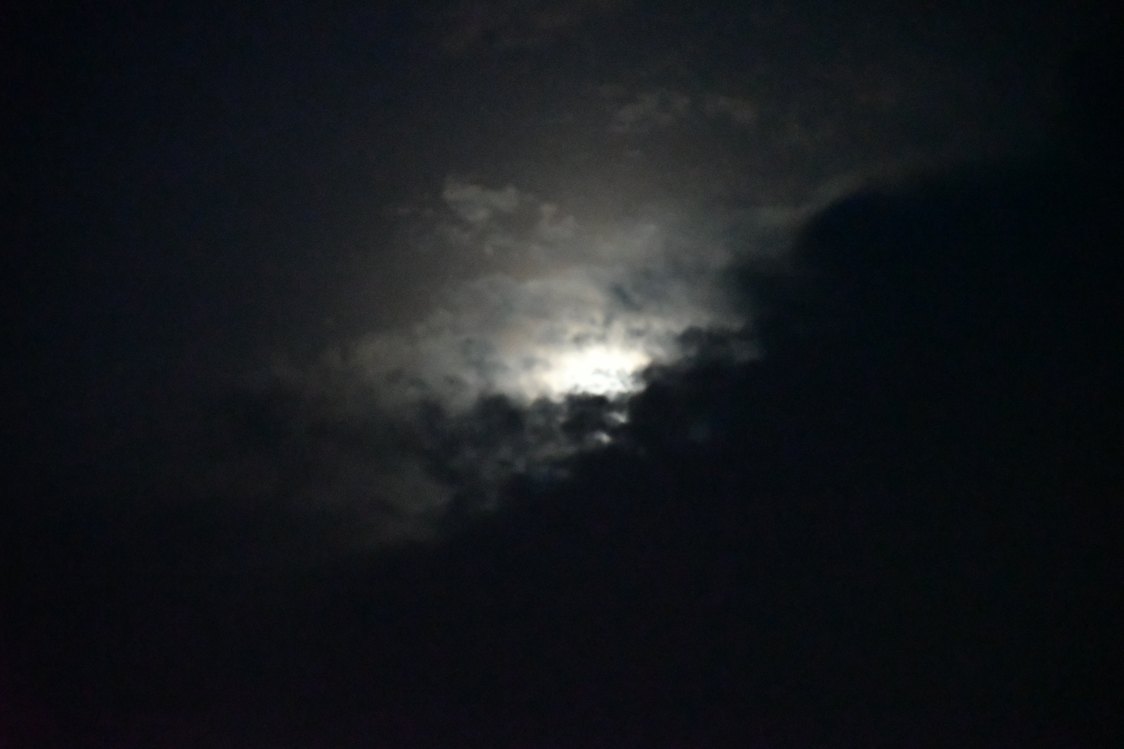 Moon partially obscured by dark clouds in night sky, bright light peeking through clouds