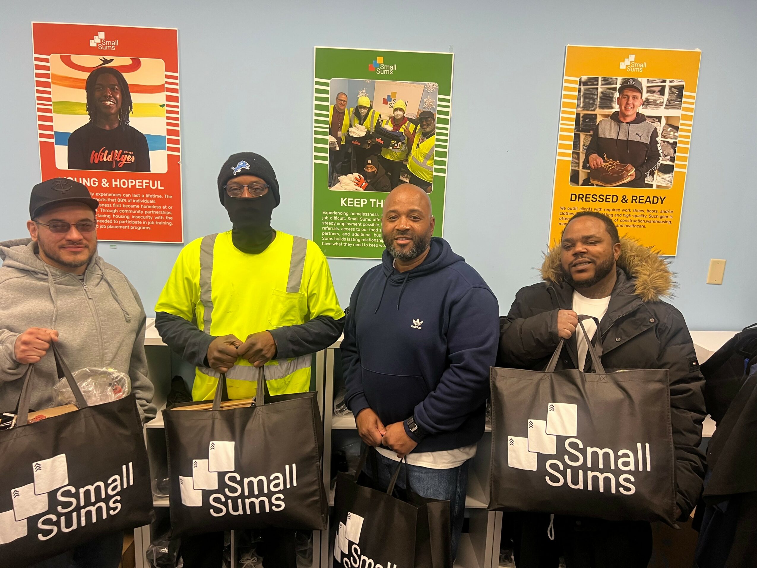 Four men standing indoors holding black bags with 'Small Sums' logo, colorful posters on the wall behind them.