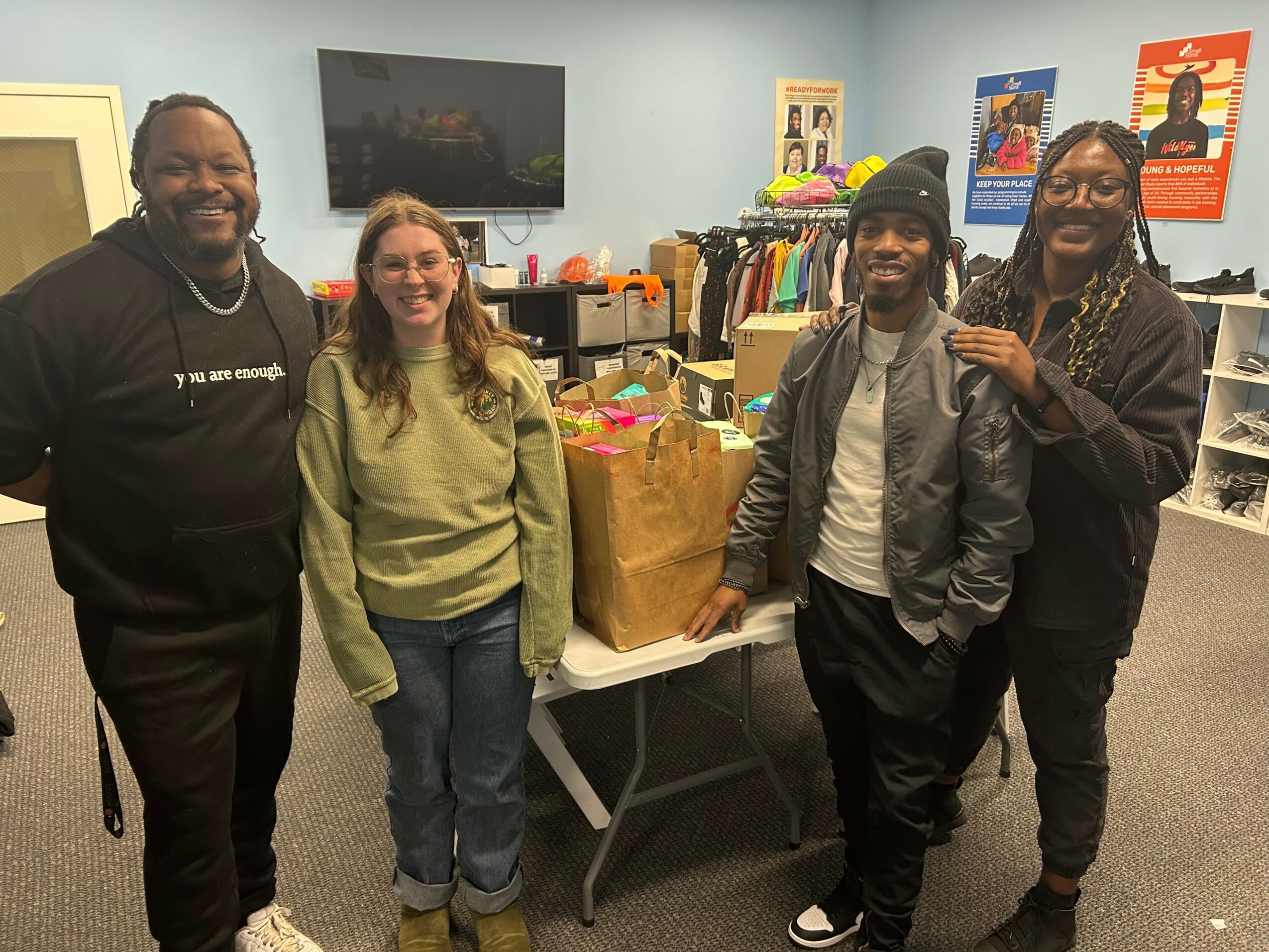 Four people standing in a room with a table of gift bags, posters, and shelves, smiling at the camera.