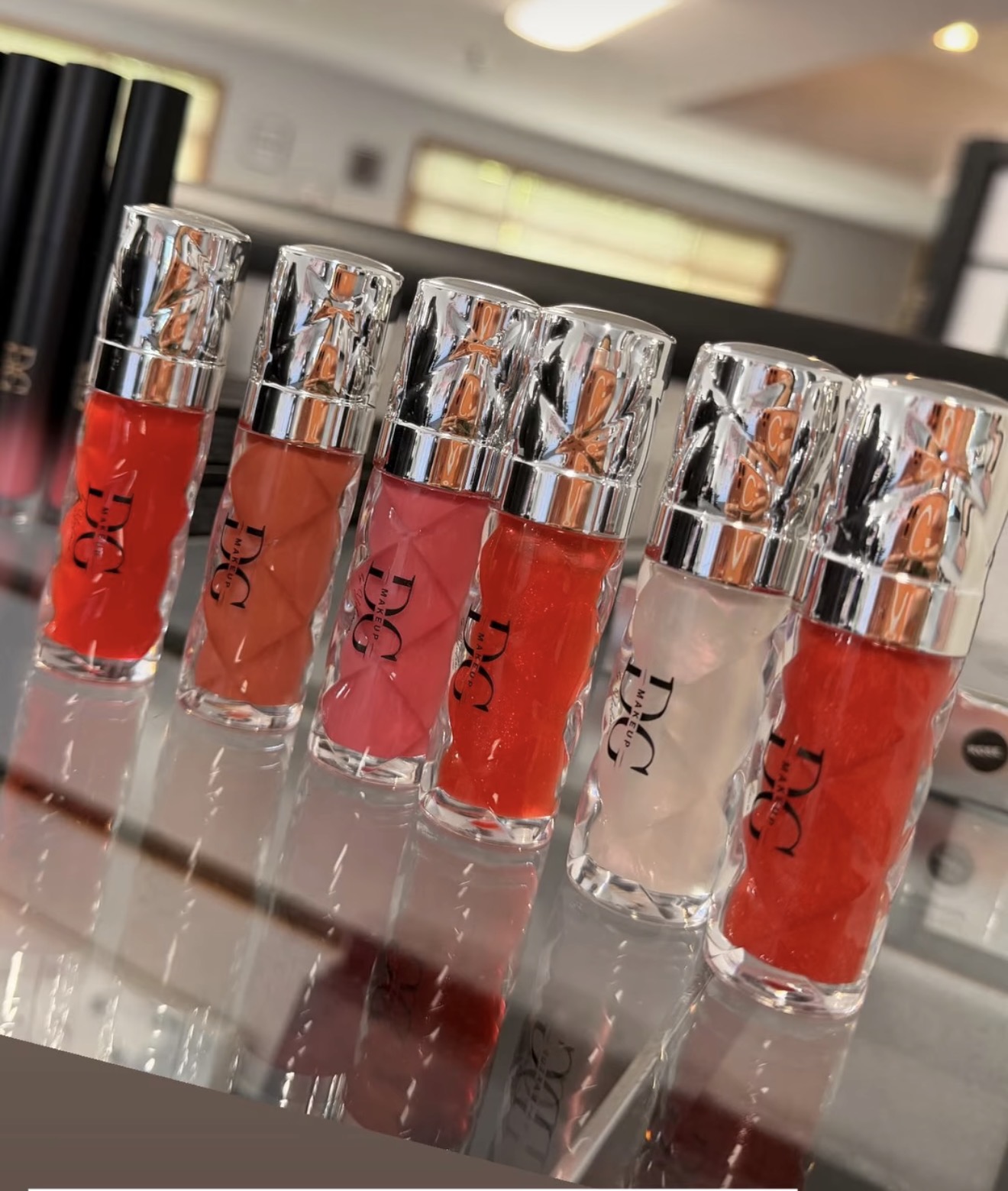Six lip gloss tubes with metallic caps arranged in a row, with a blurred background. Reflective surface beneath them.