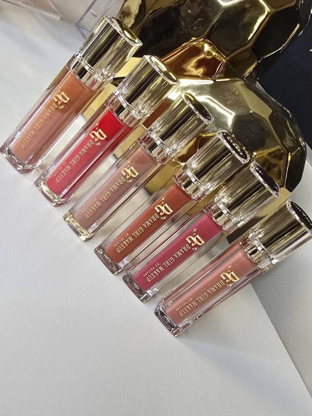 Six lip gloss tubes with gold caps arranged on a white surface, with gold decorative objects in the background.