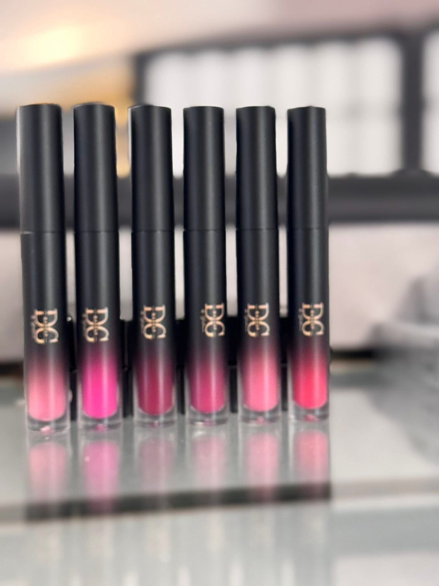 Six black and pink lip gloss tubes arranged in a row on a reflective surface, with a blurred background.