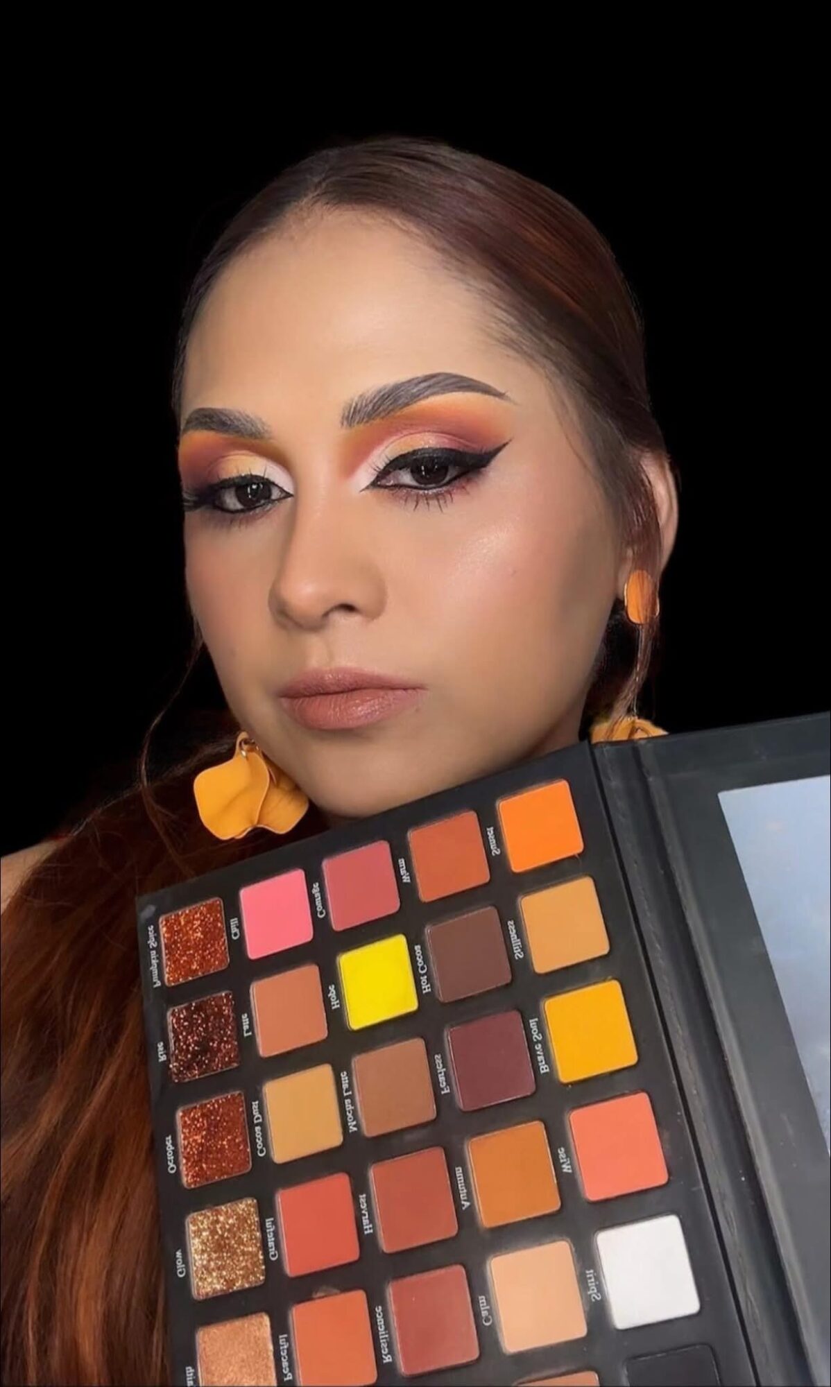 Woman with makeup holding a colorful eyeshadow palette, black background, long brown hair, orange earrings.