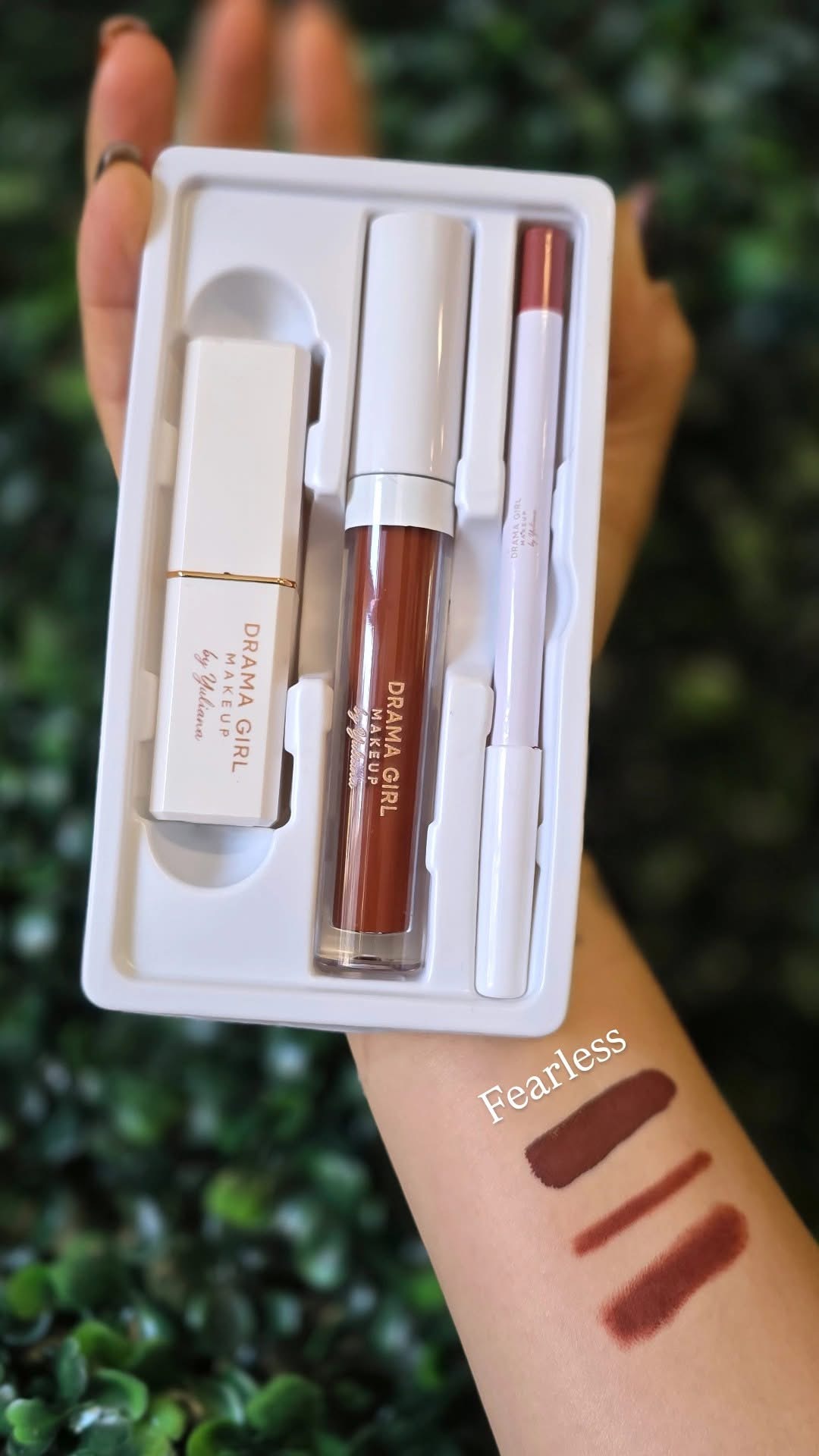 Hand holding a box with makeup products, including a lip gloss, a small brush, and swatches on arm, outdoors.