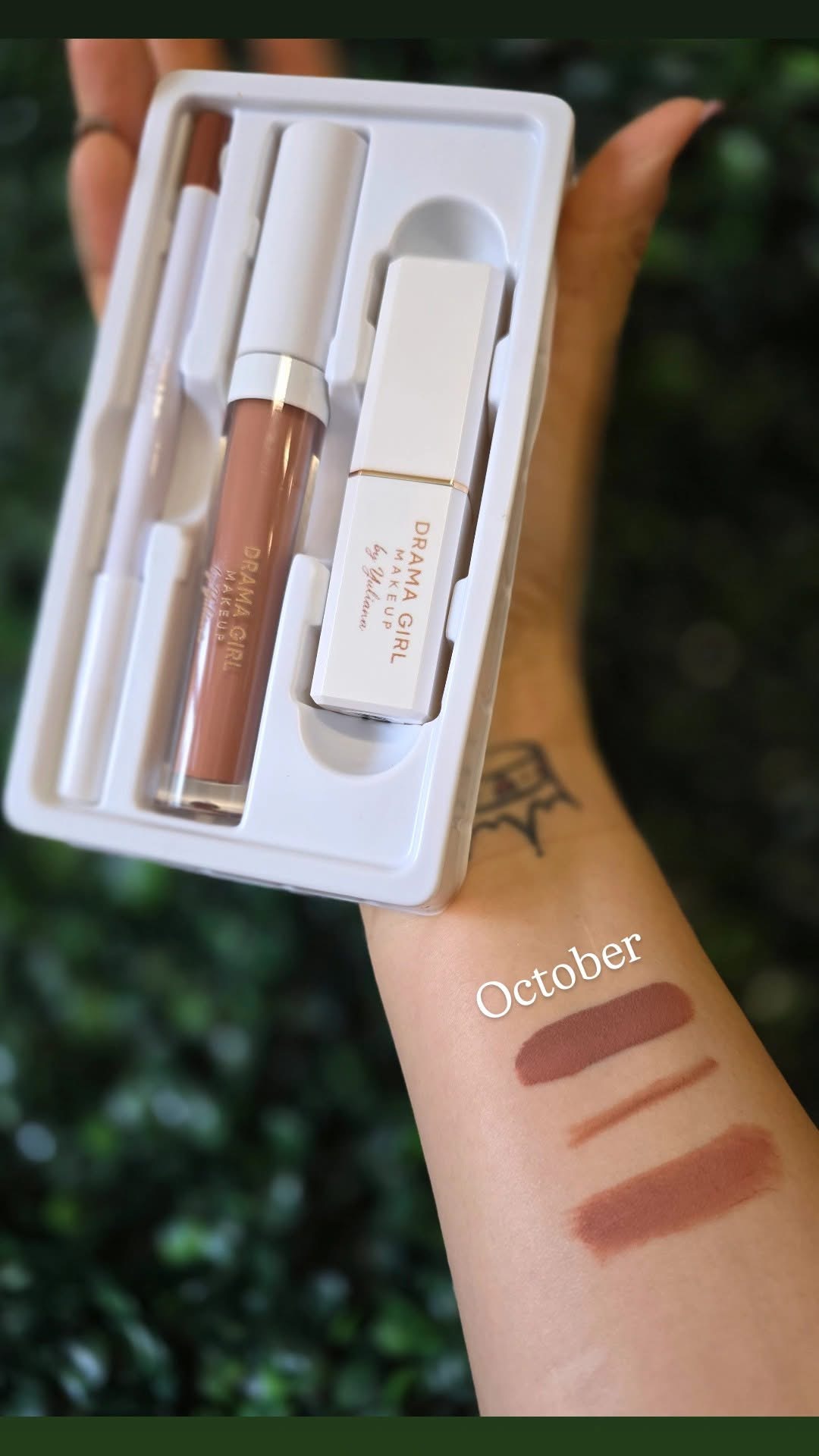 Swatch of three makeup shades on a person's arm, with a white box containing makeup tools in the background.