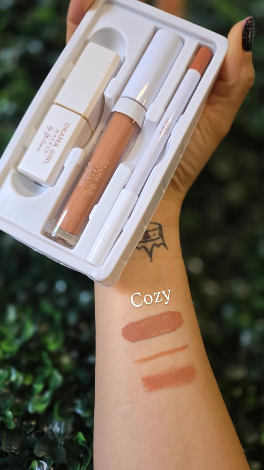 Hand holding a makeup set with a lipstick, lip gloss, and applicator, swatches on arm, outdoors background.