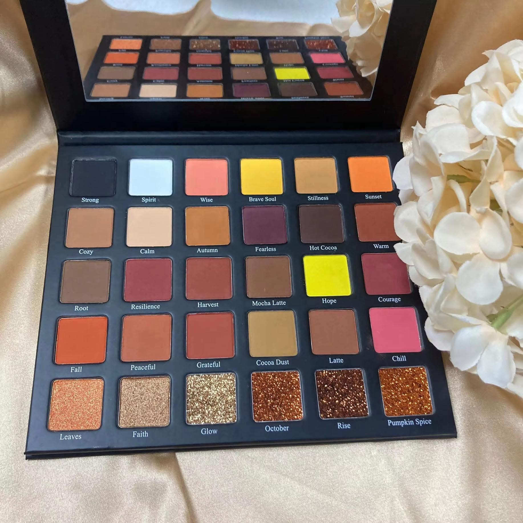Open eyeshadow palette with various shades, mirror reflecting palette, white flowers on side, on beige fabric