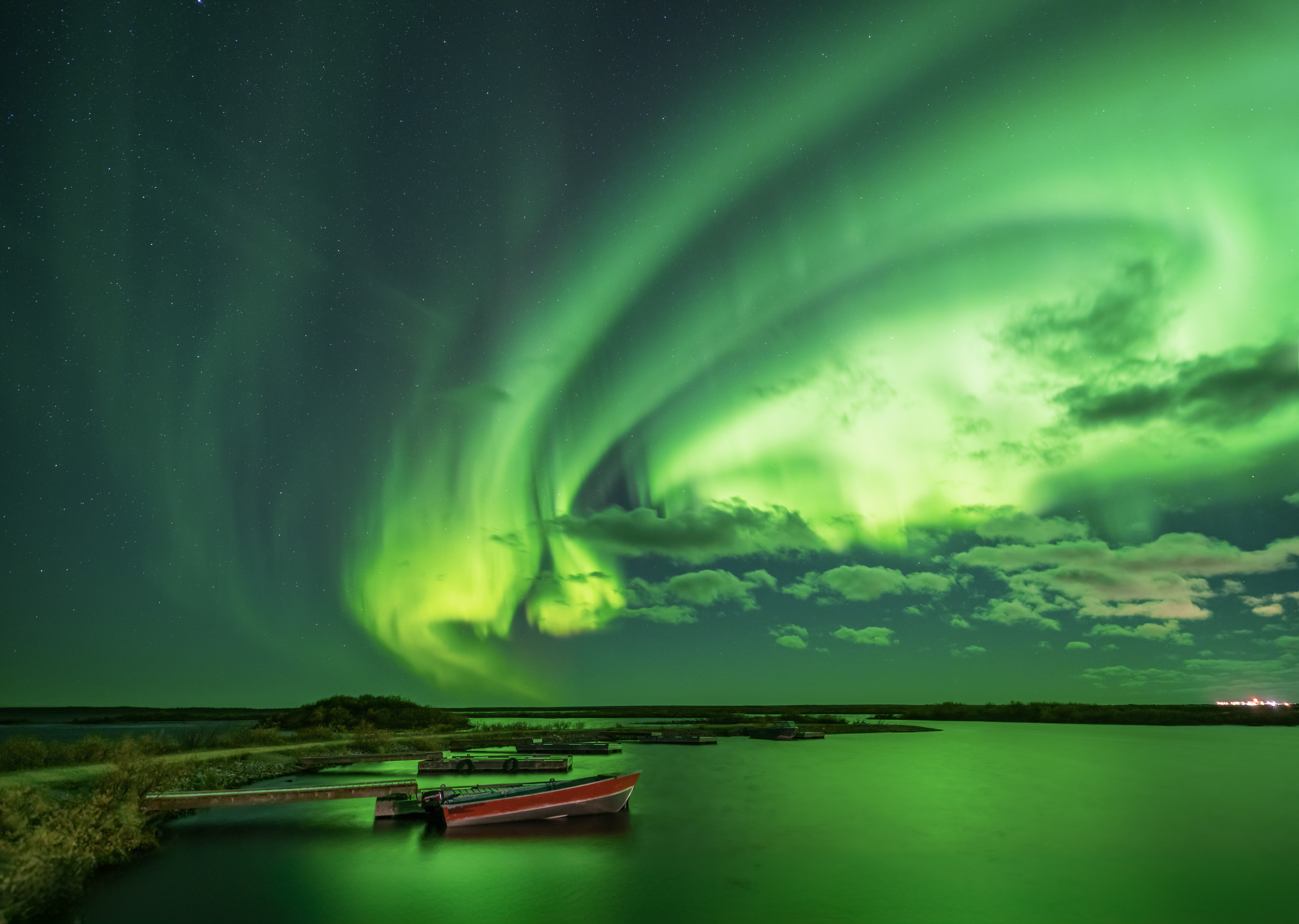 Northern lights display with green aurora swirling in the sky over a lake with boats, and a distant shoreline.